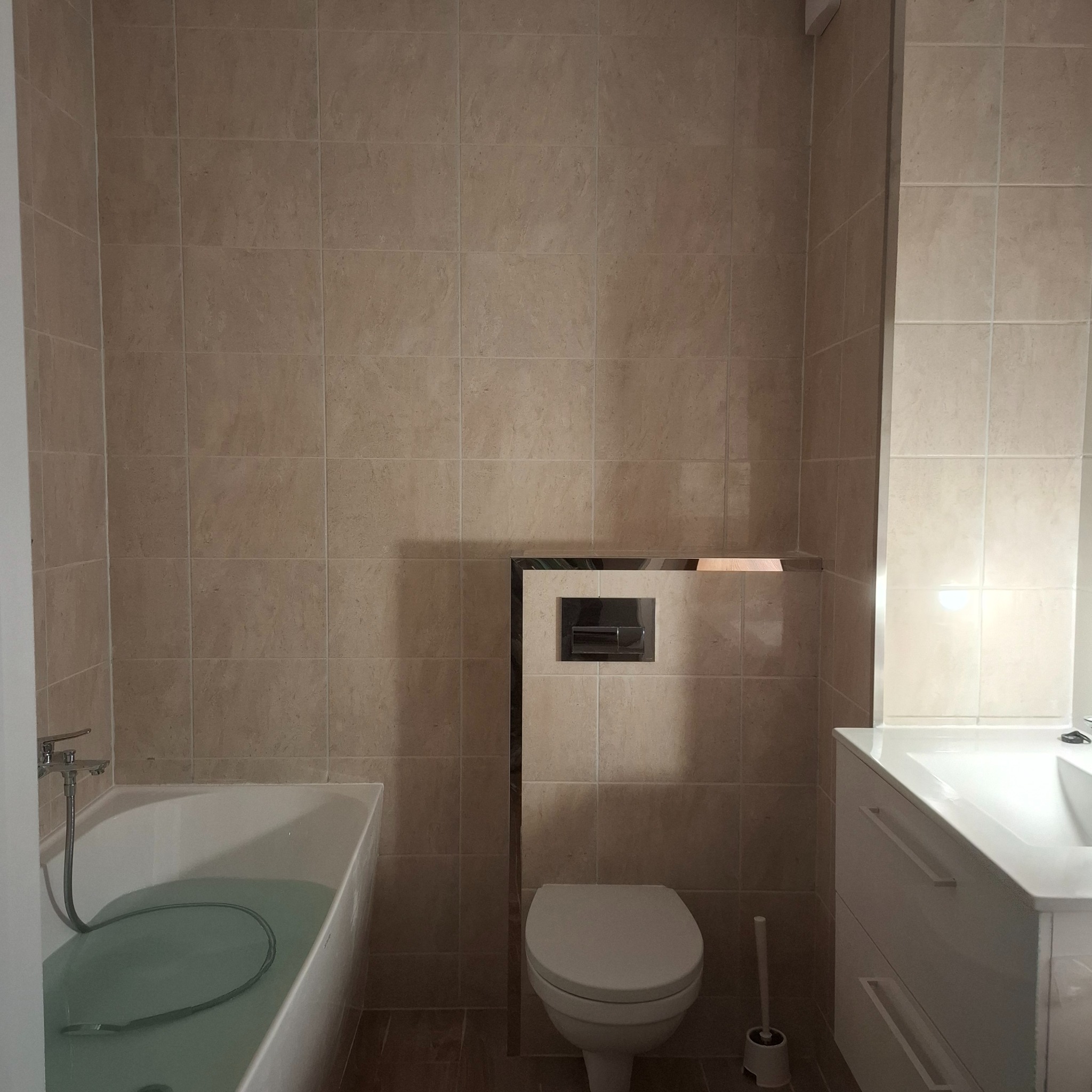 Beżowa łazienka po remoncie: płytki ścienne i podłogowe, biała wanna z wodą, toaleta podtynkowa i umywalka z szafką. Widok na całe pomieszczenie.