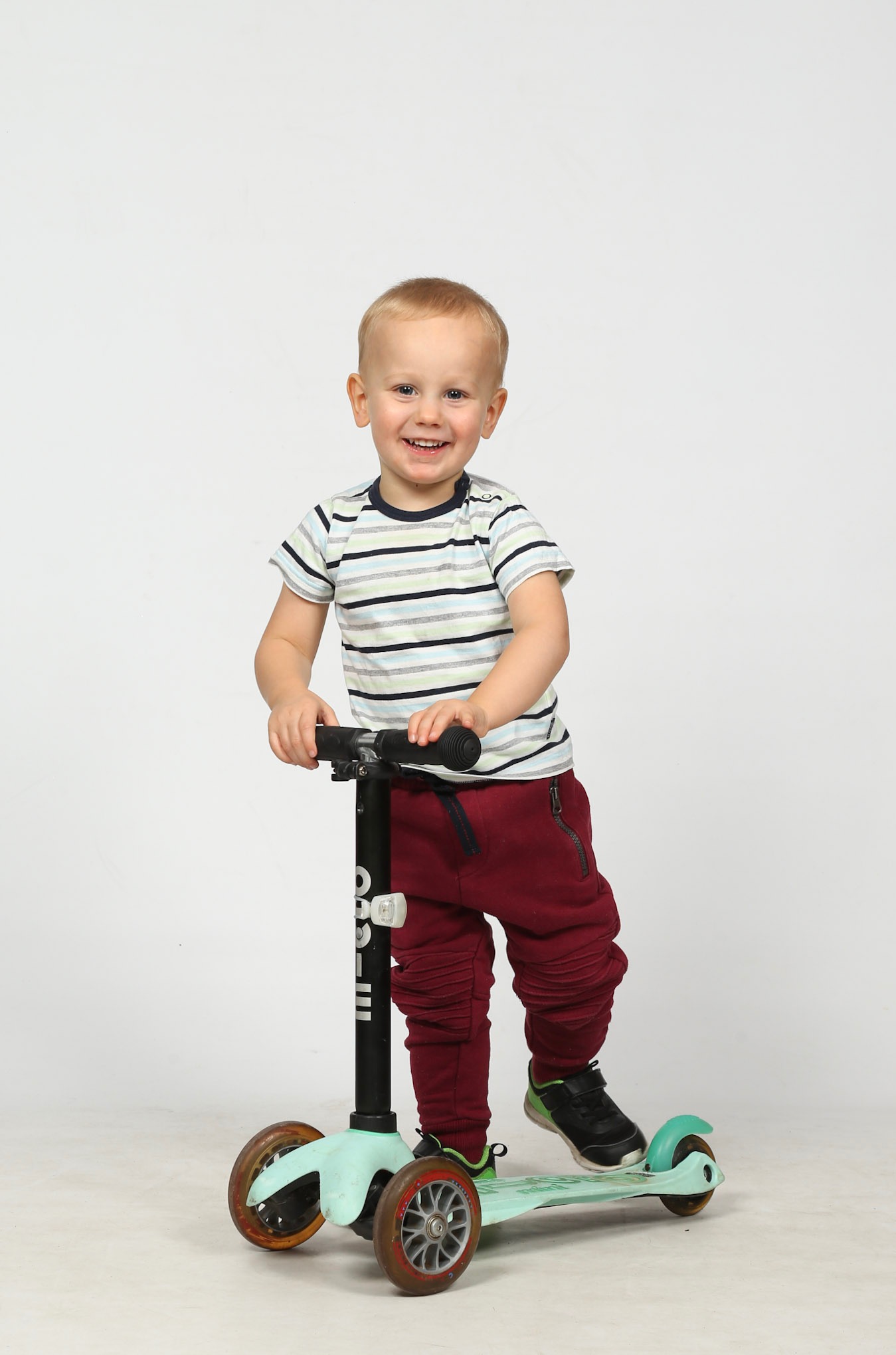 Uśmiechnięty chłopiec w paski na hulajnodze Micro Mini Deluxe, w bordowych spodniach i czarnych butach, na białym tle. Widok z przodu, centralnie.
