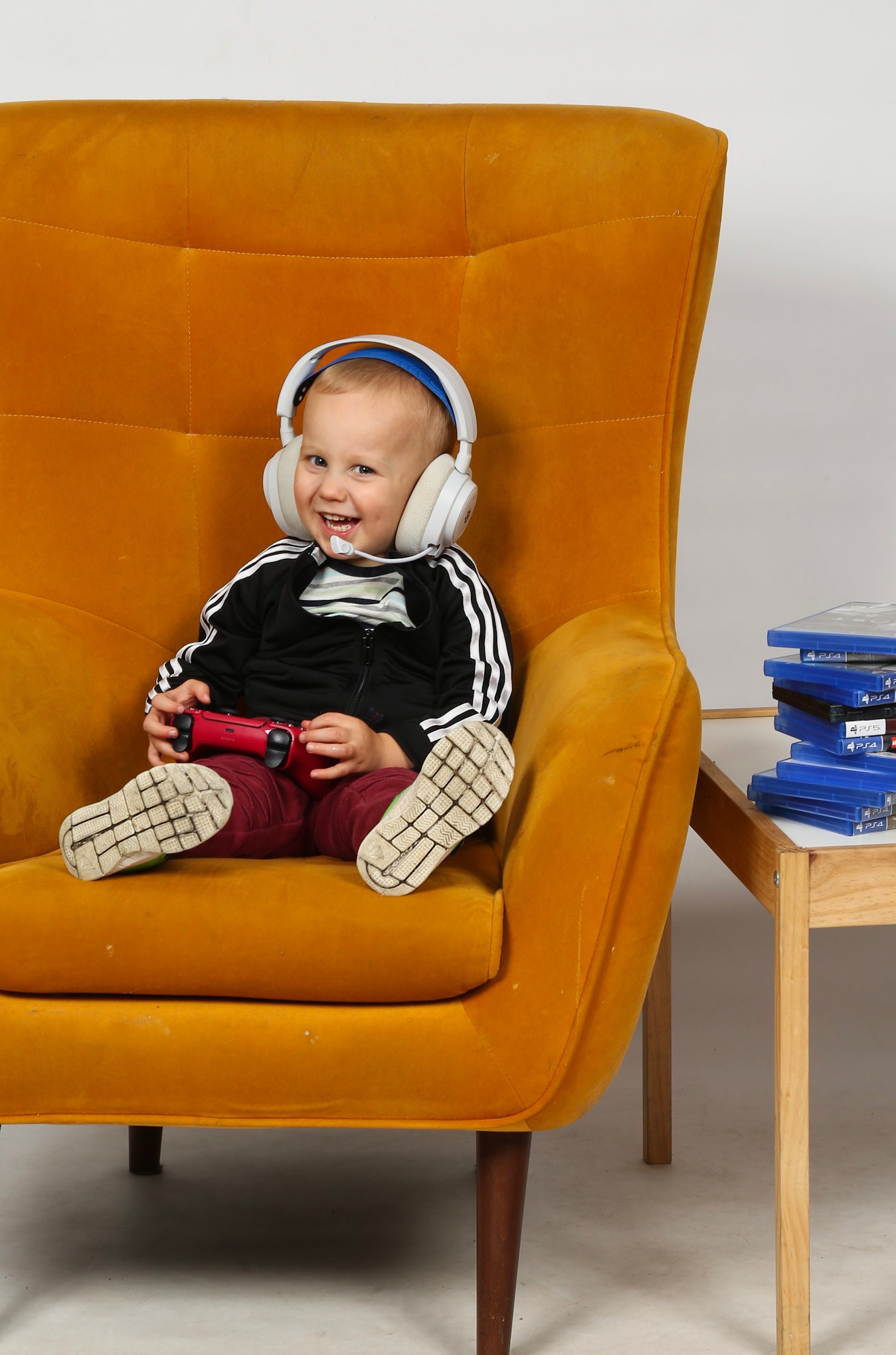 Uśmiechnięty chłopiec w słuchawkach siedzi w fotelu, trzymając czerwony kontroler do gier. Obok stolik z grami PS4. Jasne tło, naturalne światło.