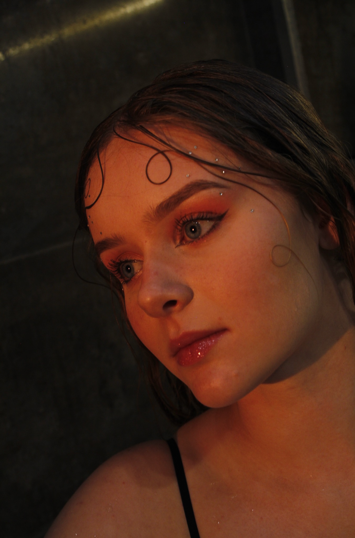 Portret młodej kobiety z artystycznym makijażem oczu i błyszczącymi ozdobami na twarzy, w ciepłym oświetleniu i z ciemnym tłem, skierowana w lewo.