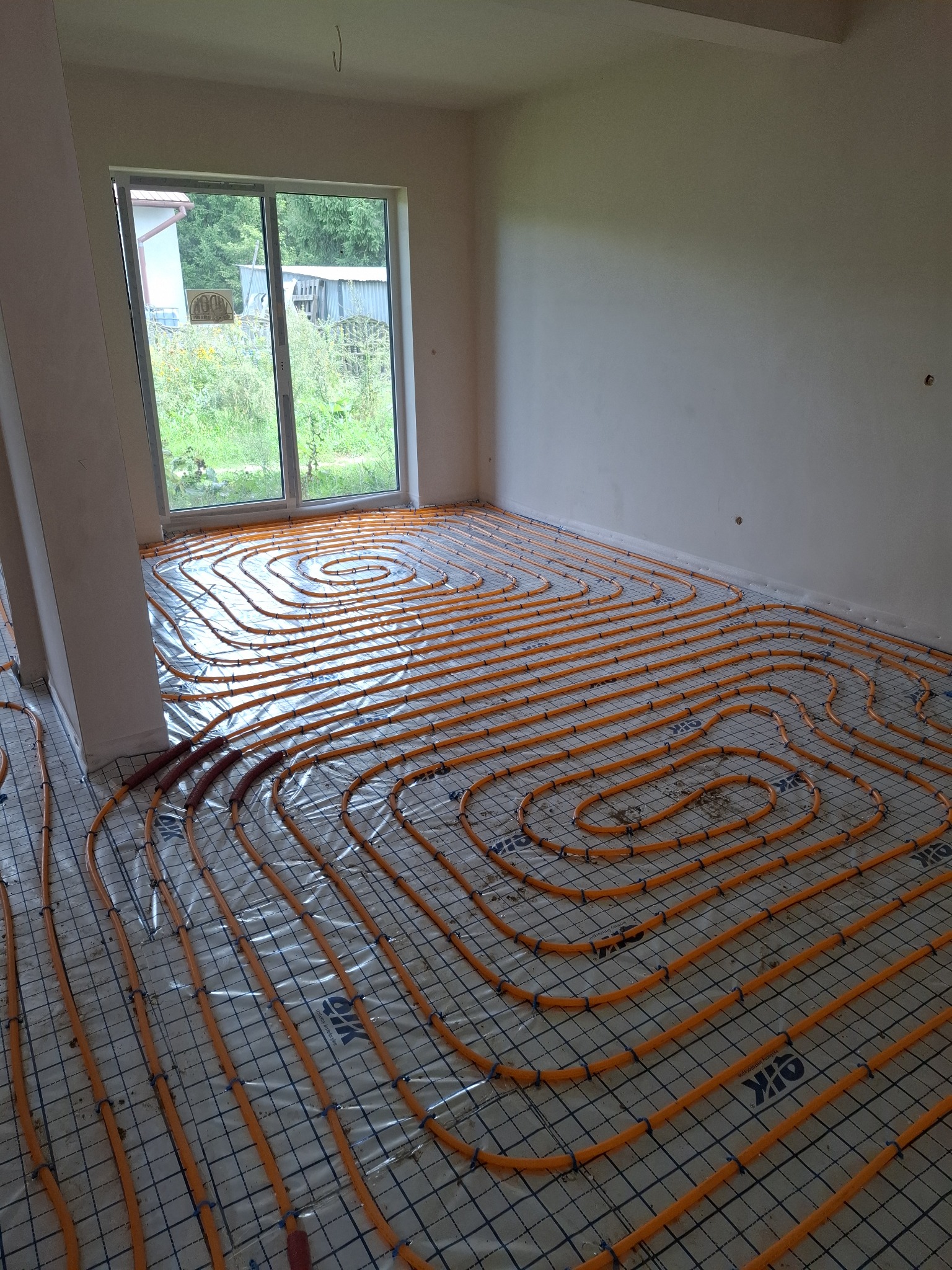 Pomarańczowe rury ogrzewania podłogowego ułożone spiralnie na folii z siatką w jasnym pokoju z dużym oknem. Widok z góry, etap instalacji przed wylewką.