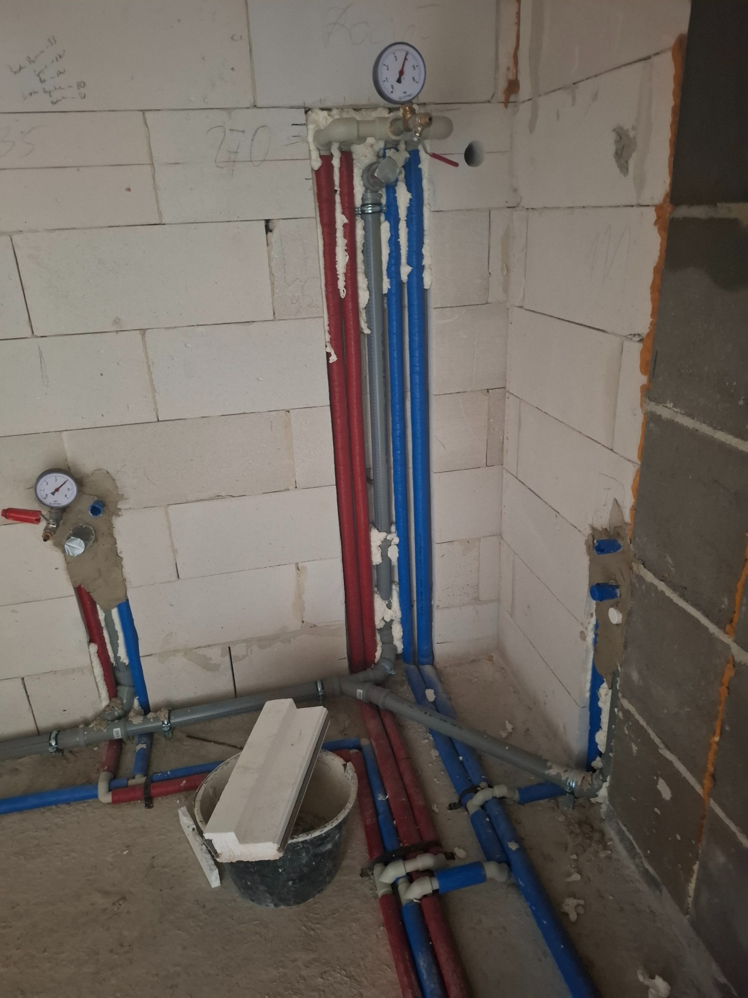 Instalacja wodna w surowym wnętrzu: czerwone i niebieskie rury prowadzone wzdłuż ścian z betonu komórkowego, manometry, pianka montażowa, wiadro z resztkami materiałów.