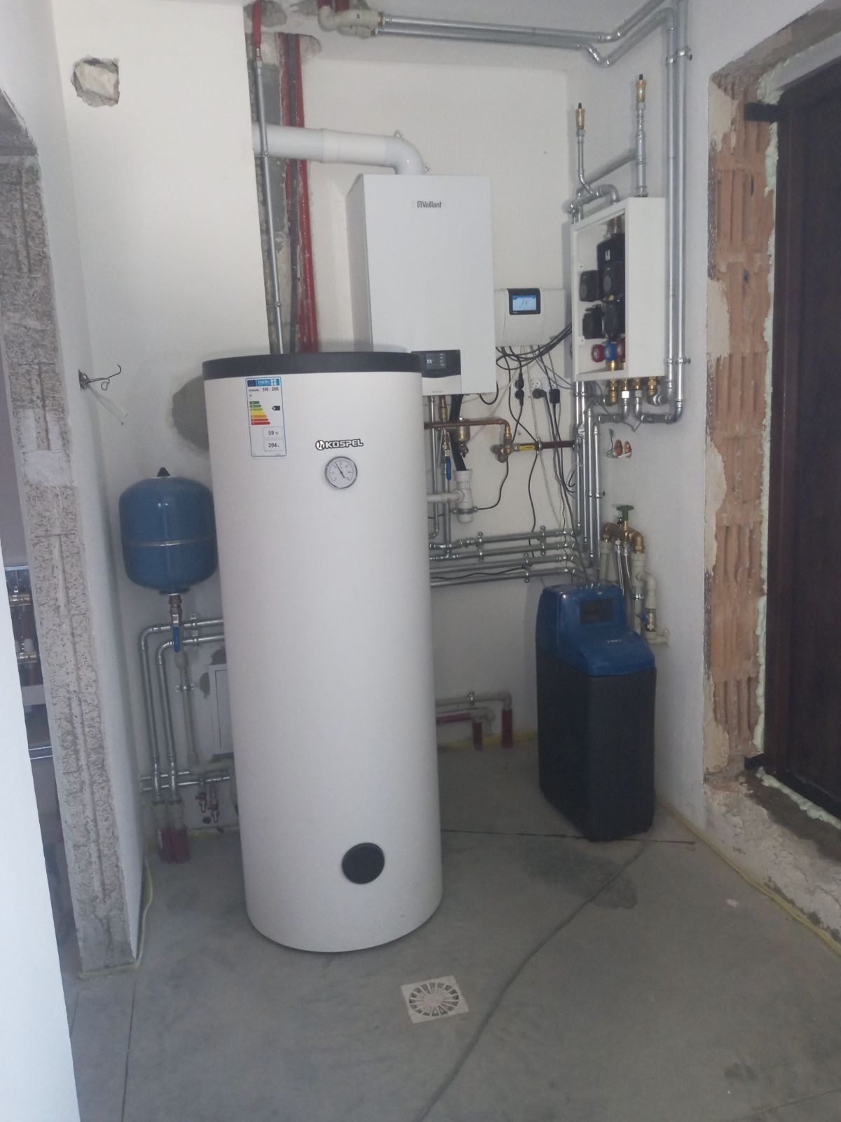 Nowoczesna kotłownia z białym zasobnikiem Kospel, piecem Vaillant i zmiękczaczem wody. Widoczne rury instalacji grzewczej i wodnej. Świeżo wykończone pomieszczenie.