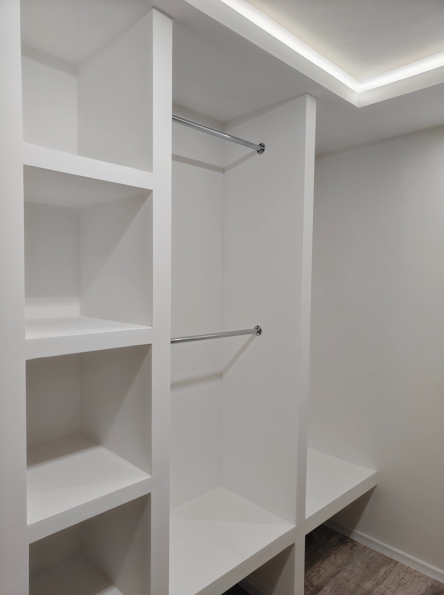 Biała, wnękowa garderoba z półkami i drążkami na ubrania, oświetlona ledowym światłem sufitowym. Minimalistyczny design, zabudowa na wymiar.