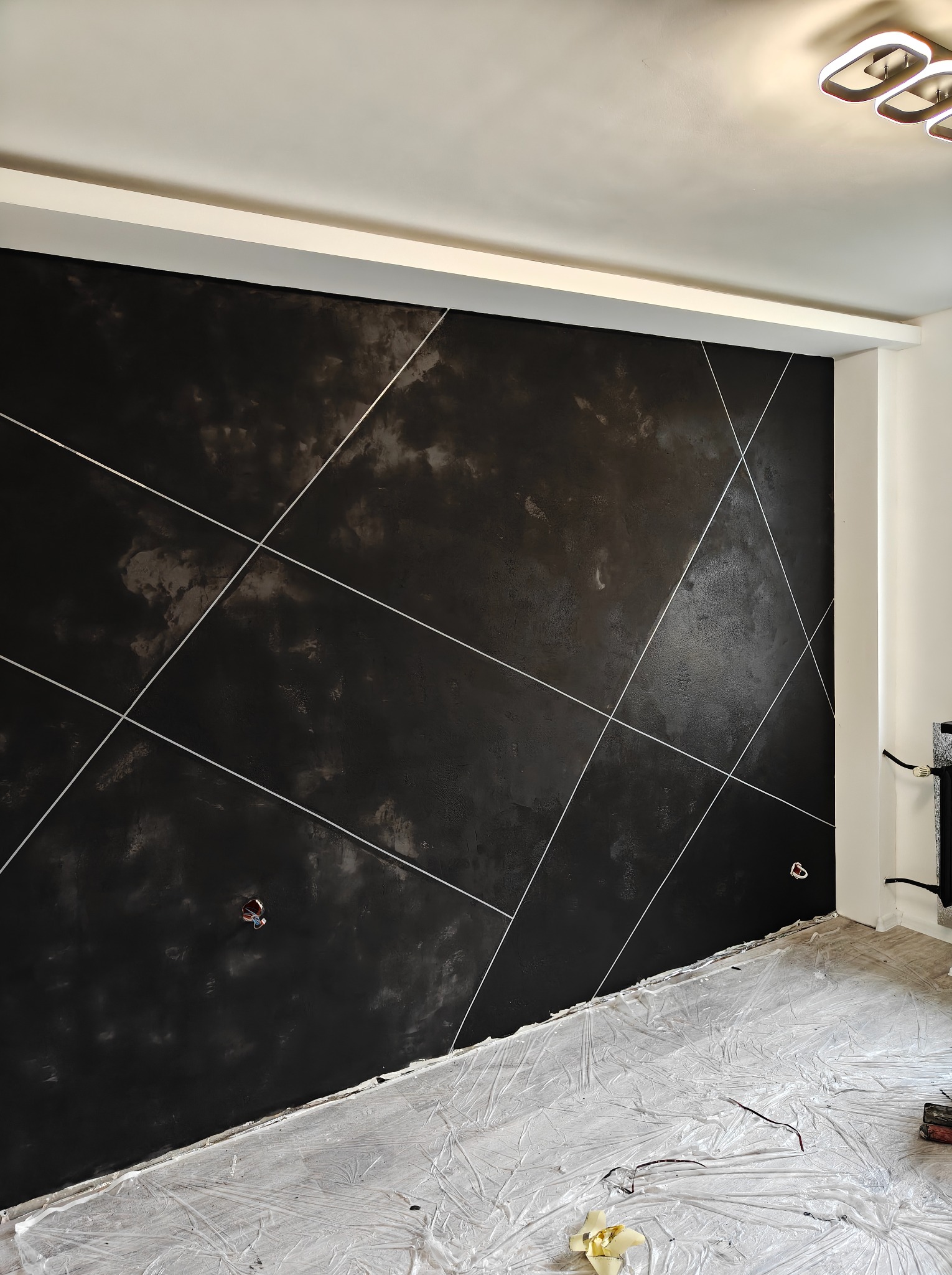 Czarna ściana z dekoracyjną fakturą i białymi liniami, zabezpieczona folią podłoga, widoczne gniazdka elektryczne. Nowoczesne wykończenie wnętrza w trakcie prac.