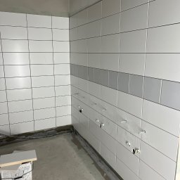 Mix-Bud - Ściana wyłożona białymi, prostokątnymi płytkami z szarym pasem dekoracyjnym. Widoczne otwory na instalacje i wystające kołki. Na podłodze resztki materiałów i pudełko z płytkami.