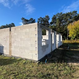 Mix-Bud - Surowy stan domu z betonu komórkowego w trakcie budowy, widoczne ściany i otwory okienne, na tle zieleni i błękitnego nieba. Palety z bloczkami obok.