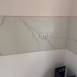 Mix-Bud - Wykończona ściana z marmurowymi płytkami i podwójnym gniazdkiem elektrycznym. Widoczne łączenie płytek z pomalowaną ścianą. Fragment narożnika pomieszczenia.