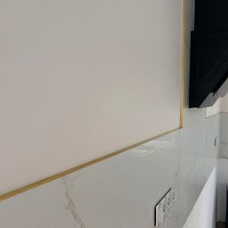 Mix-Bud - Biała ściana z dekoracyjną, złotą listwą i marmurowymi płytkami. Widoczny czarny okap kuchenny i gniazdka elektryczne. Ukośne ujęcie podkreśla detale wykończenia.