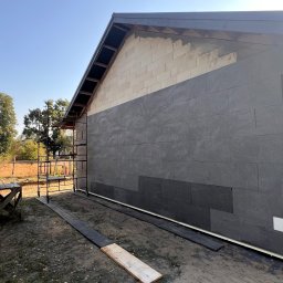 Mix-Bud - Ocieplanie elewacji domu jednorodzinnego z widocznymi fragmentami bloczków i szarymi płytami izolacyjnymi. Na pierwszym planie fragment sylwetki osoby w czapce.