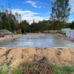 Mix-Bud - Świeżo wylana, mokra płyta fundamentowa na gruncie, otoczona piaskiem i deskowaniem. W tle składowane bloczki Solbet i zieleń drzew.