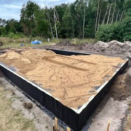 Mix-Bud - Przygotowany wykop pod fundament z izolacją pionową i zasypany piaskiem, widoczne ślady opon. W tle drzewa i sprzęt budowlany.