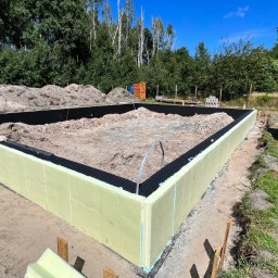 Mix-Bud - Ocieplone fundamenty domu jednorodzinnego z widocznymi bloczkami Solbet w tle, na tle zieleni i błękitnego nieba. Etap budowy w słoneczny dzień.