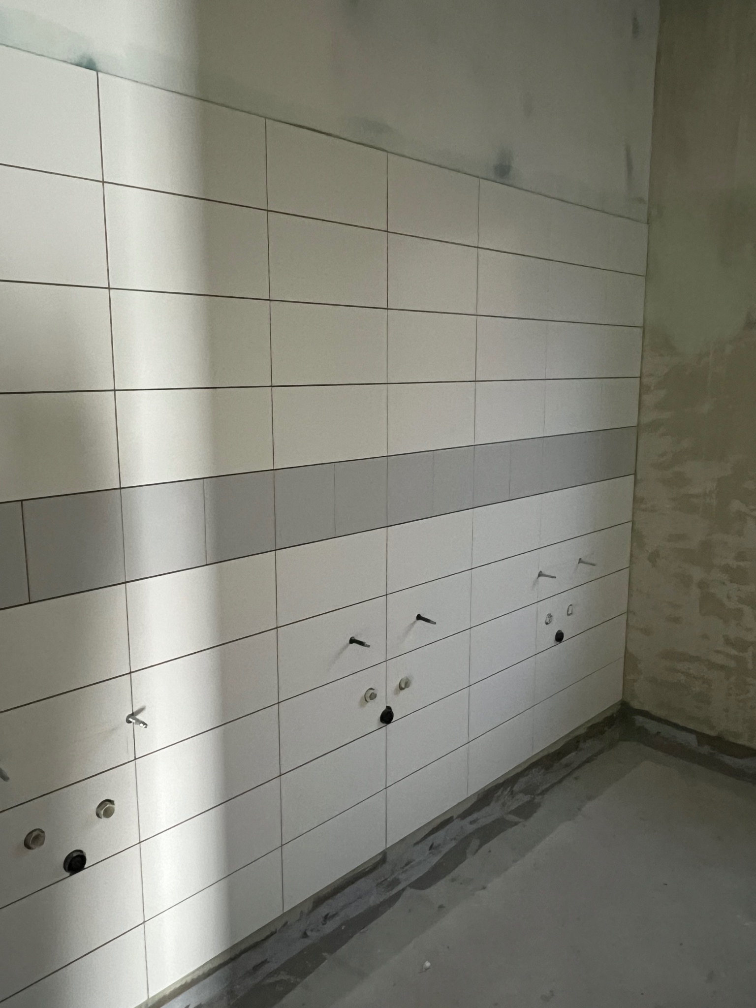 Ściana wyłożona białymi, prostokątnymi płytkami z szarą linią dekoracyjną. Widoczne otwory na instalacje elektryczne i wodne. Surowy stan wykończenia wnętrza.