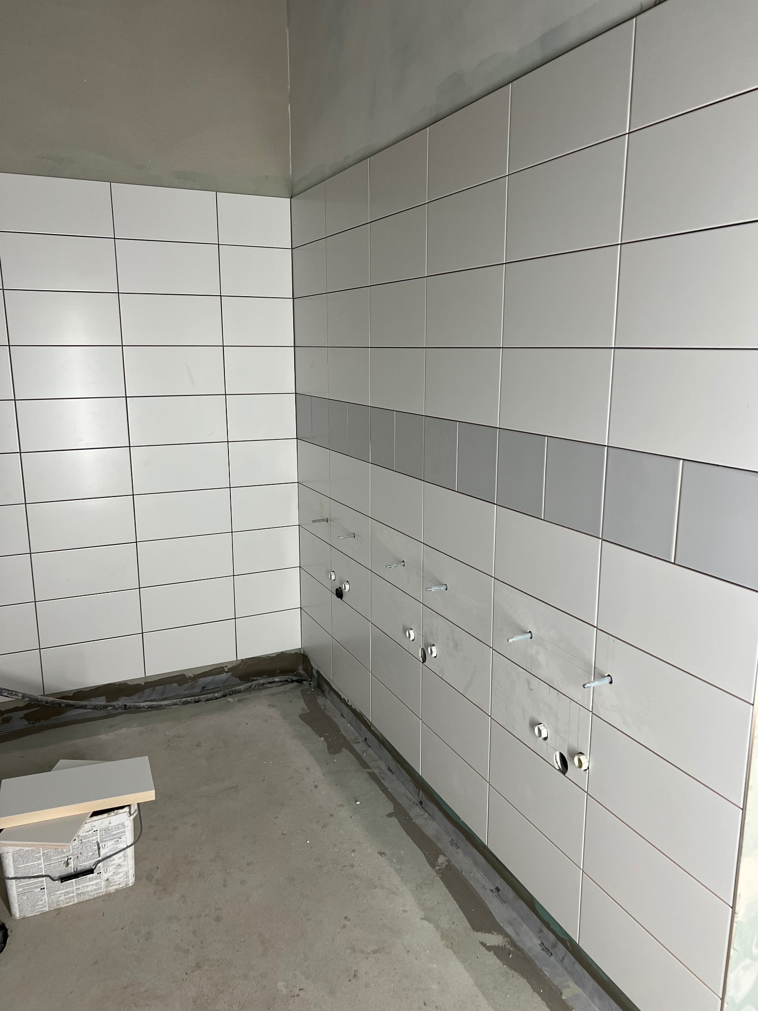 Ściana wyłożona białymi, prostokątnymi płytkami z szarym pasem dekoracyjnym. Widoczne otwory na instalacje i wystające kołki. Na podłodze resztki materiałów i pudełko z płytkami.