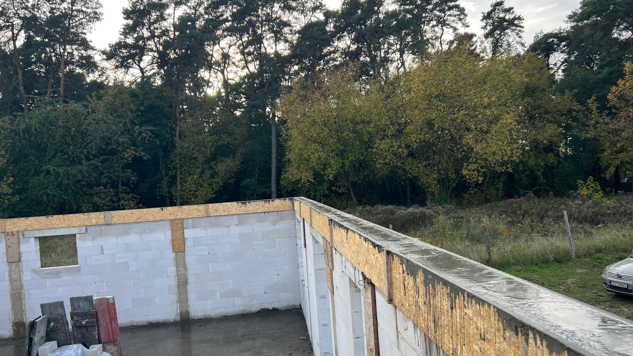 Wznoszenie ścian z betonu komórkowego na budowie domu jednorodzinnego. Widoczne nadproża okienne i zbrojenie wieńca. W tle drzewa i fragment samochodu.