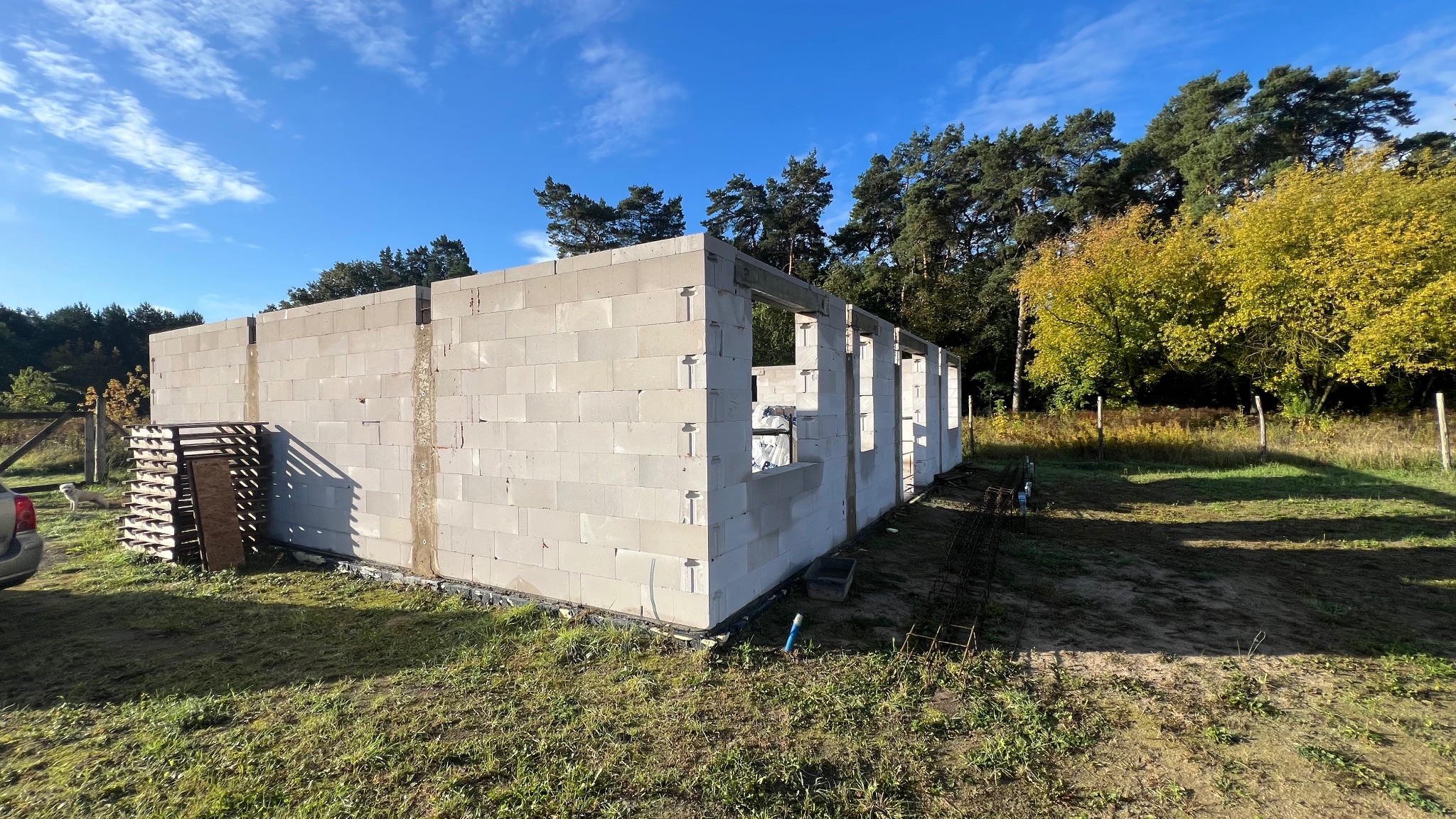 Surowy stan domu z betonu komórkowego w trakcie budowy, widoczne ściany i otwory okienne, na tle zieleni i błękitnego nieba. Palety z bloczkami obok.