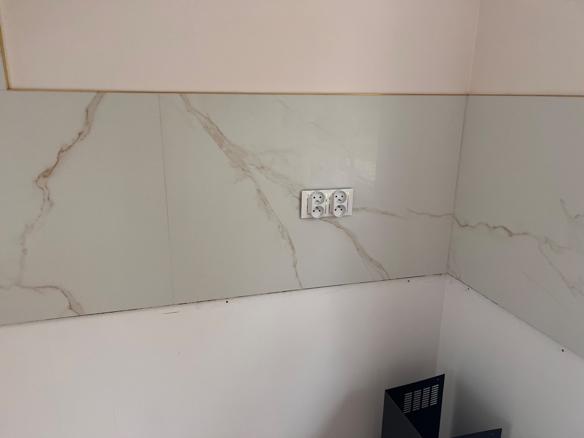 Wykończona ściana z marmurowymi płytkami i podwójnym gniazdkiem elektrycznym. Widoczne łączenie płytek z pomalowaną ścianą. Fragment narożnika pomieszczenia.