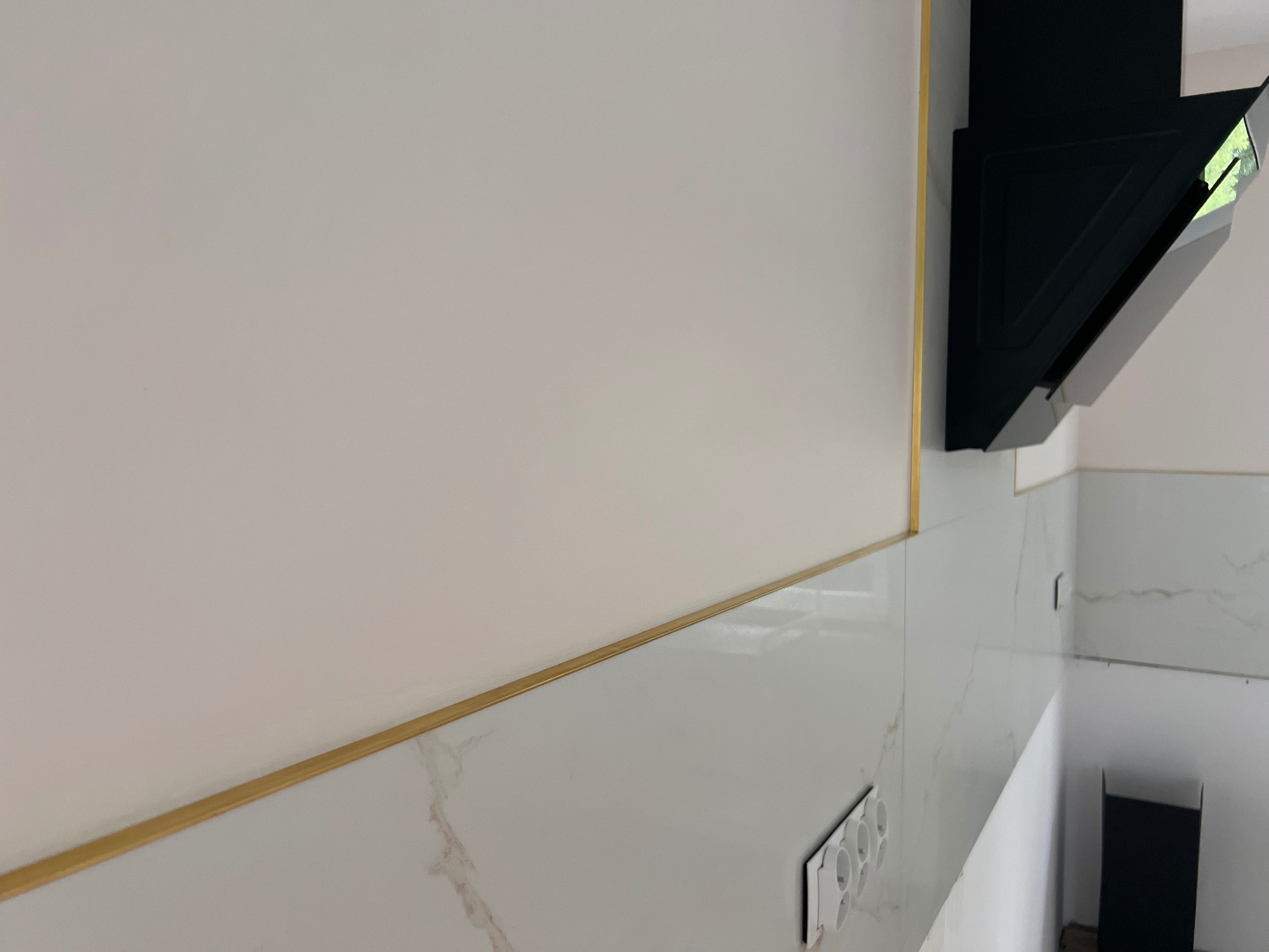 Biała ściana z dekoracyjną, złotą listwą i marmurowymi płytkami. Widoczny czarny okap kuchenny i gniazdka elektryczne. Ukośne ujęcie podkreśla detale wykończenia.
