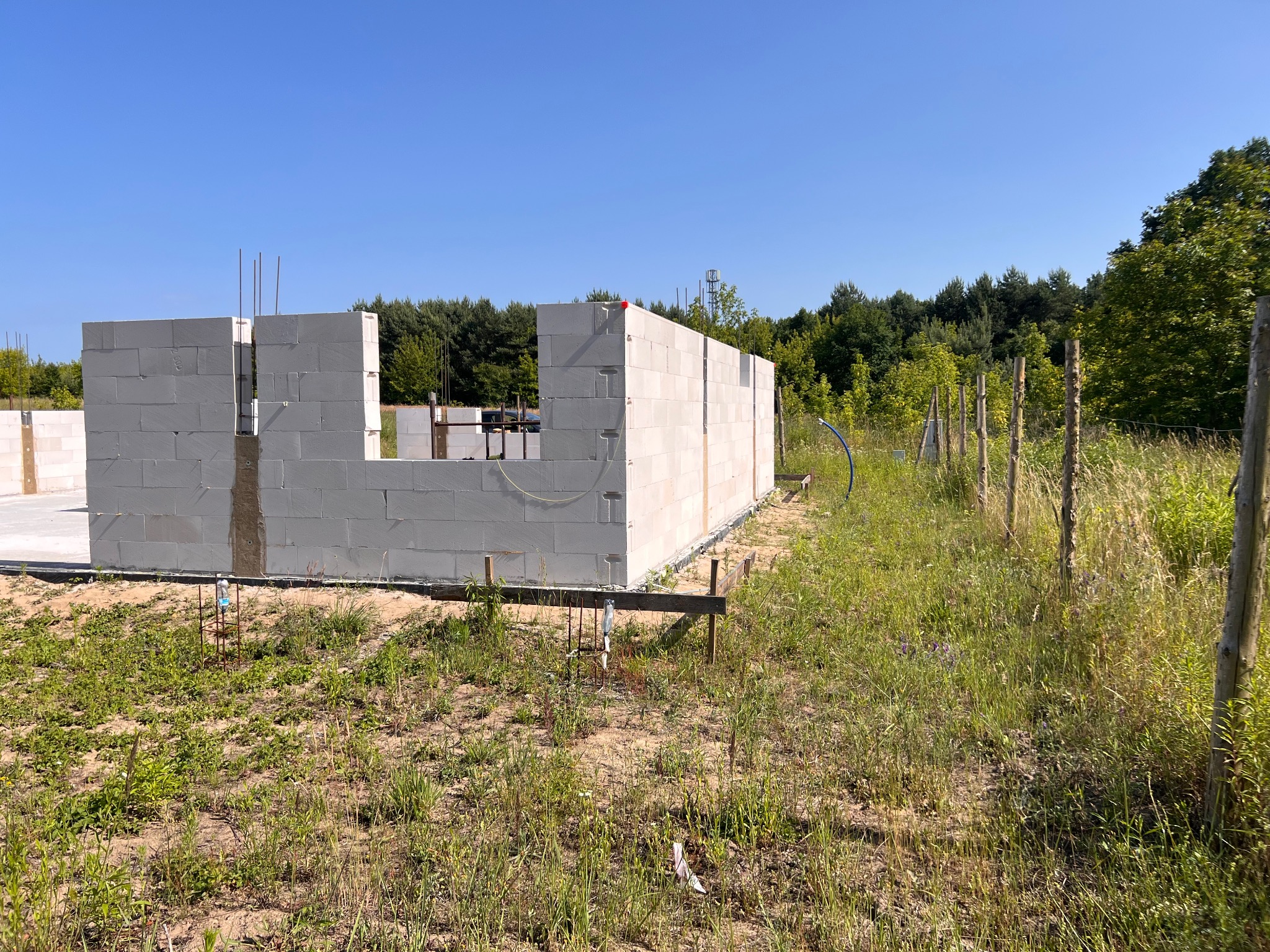 Wznoszone ściany z białych bloczków betonu komórkowego na fundamencie, widoczne pręty zbrojeniowe, w tle zieleń drzew i błękitne niebo.