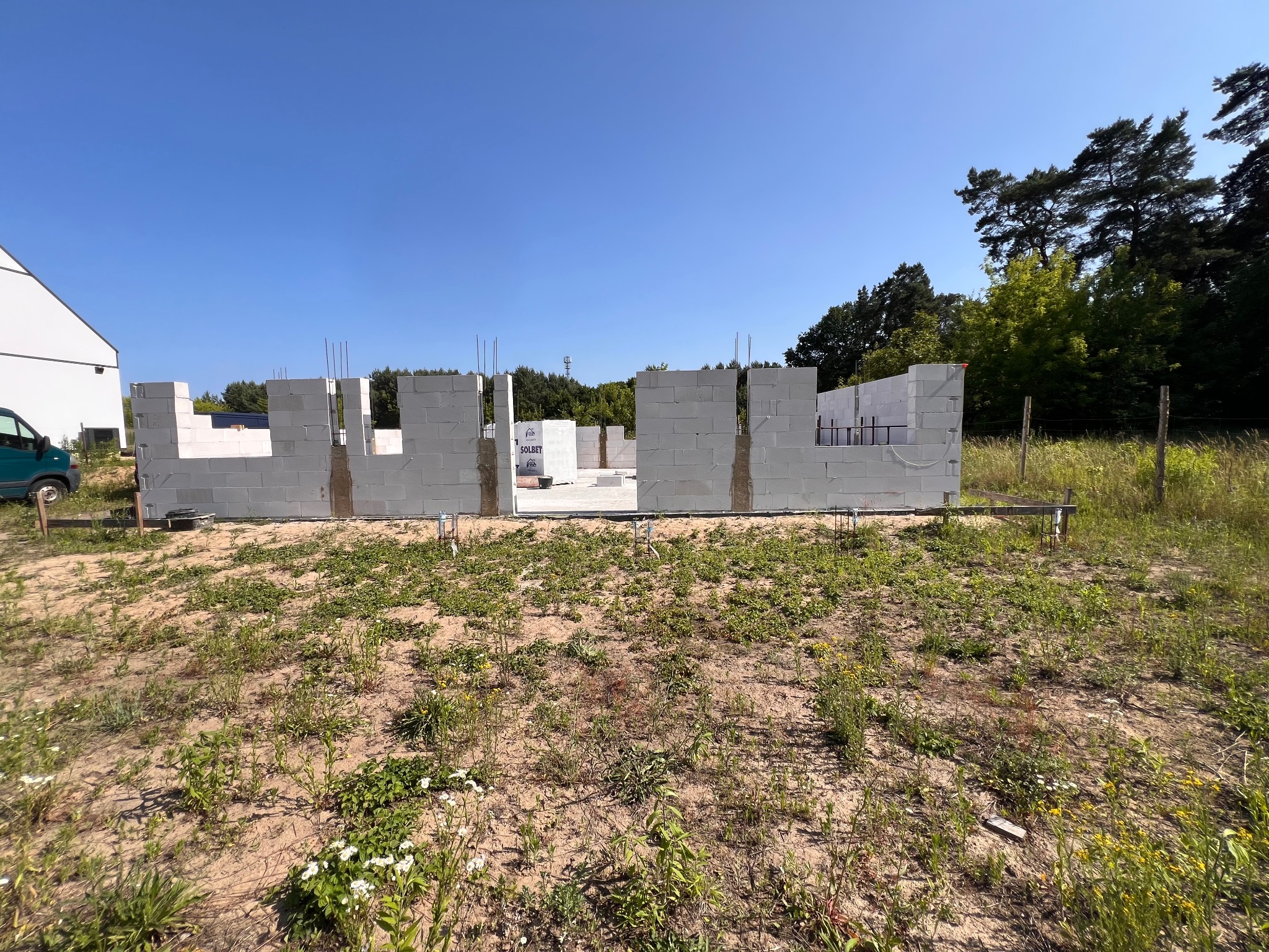 Wznoszone ściany domu z białych bloczków Solbet, widoczne zbrojenia pionowe, w tle zieleń drzew i błękitne niebo. Budowa w toku, widok z poziomu gruntu.