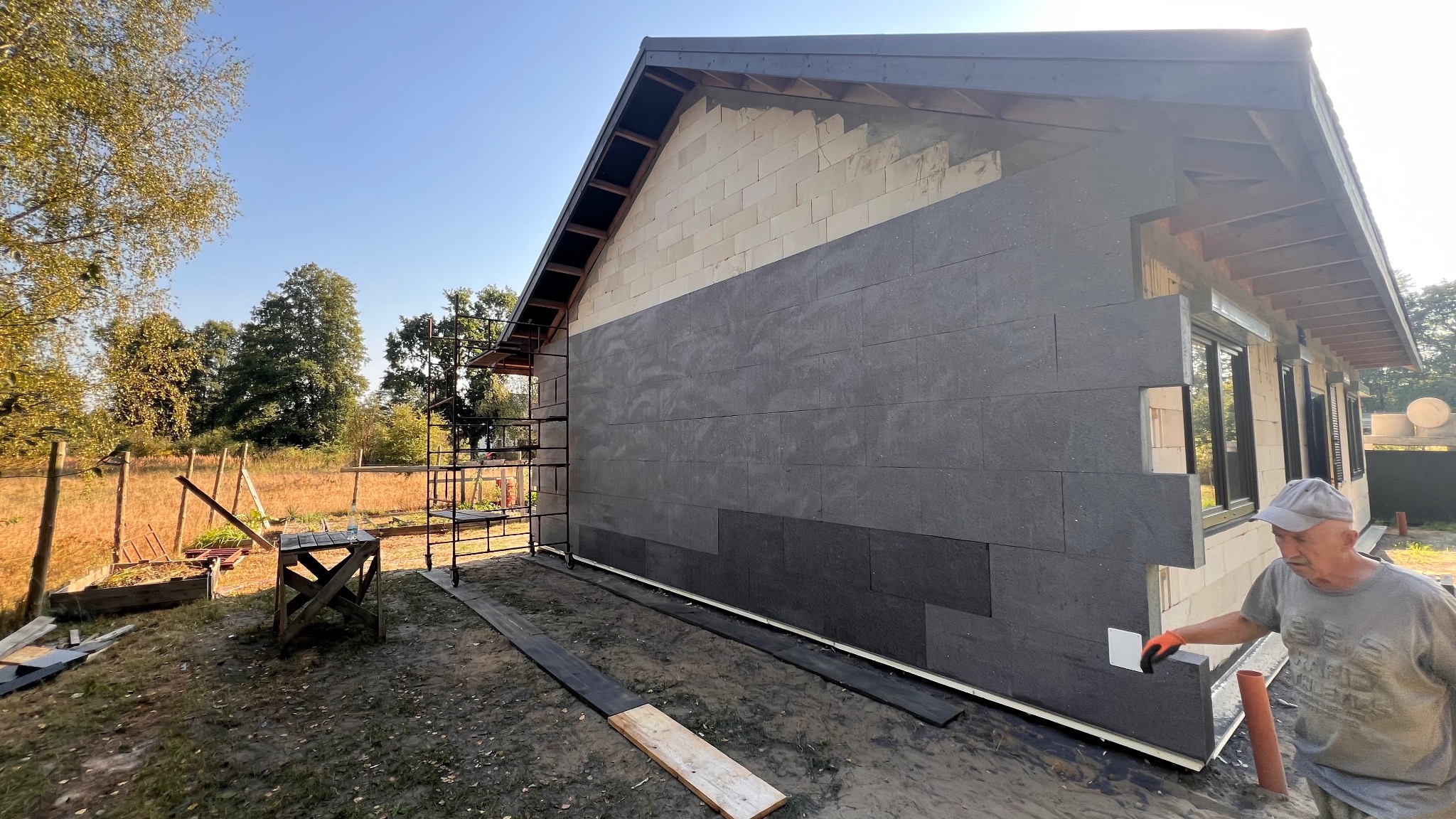 Ocieplanie elewacji domu jednorodzinnego z widocznymi fragmentami bloczków i szarymi płytami izolacyjnymi. Na pierwszym planie fragment sylwetki osoby w czapce.