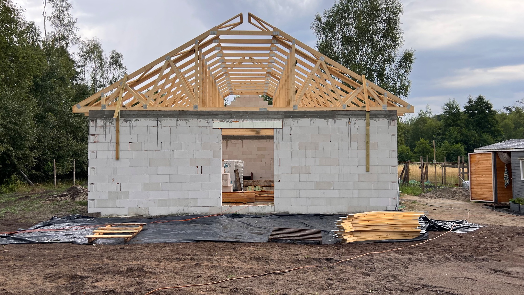 Budowa domu z białych bloczków: ściany wzniesione, drewniana konstrukcja dachu w trakcie montażu, widoczne materiały budowlane. Etap surowy otwarty.