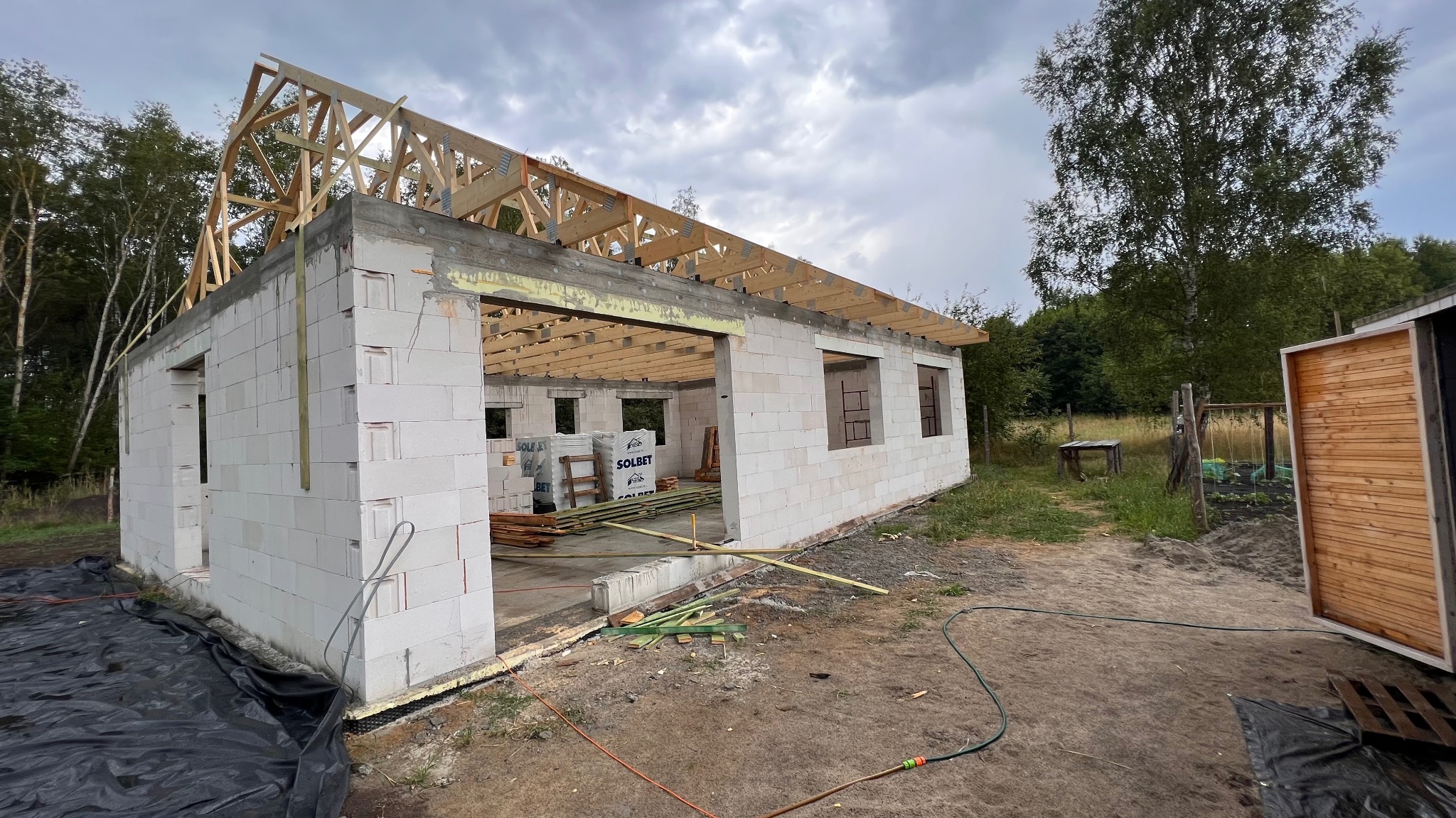 Budowa domu jednorodzinnego: ściany z betonu komórkowego, drewniana konstrukcja dachu w trakcie montażu. Widoczne materiały budowlane i otoczenie placu budowy.