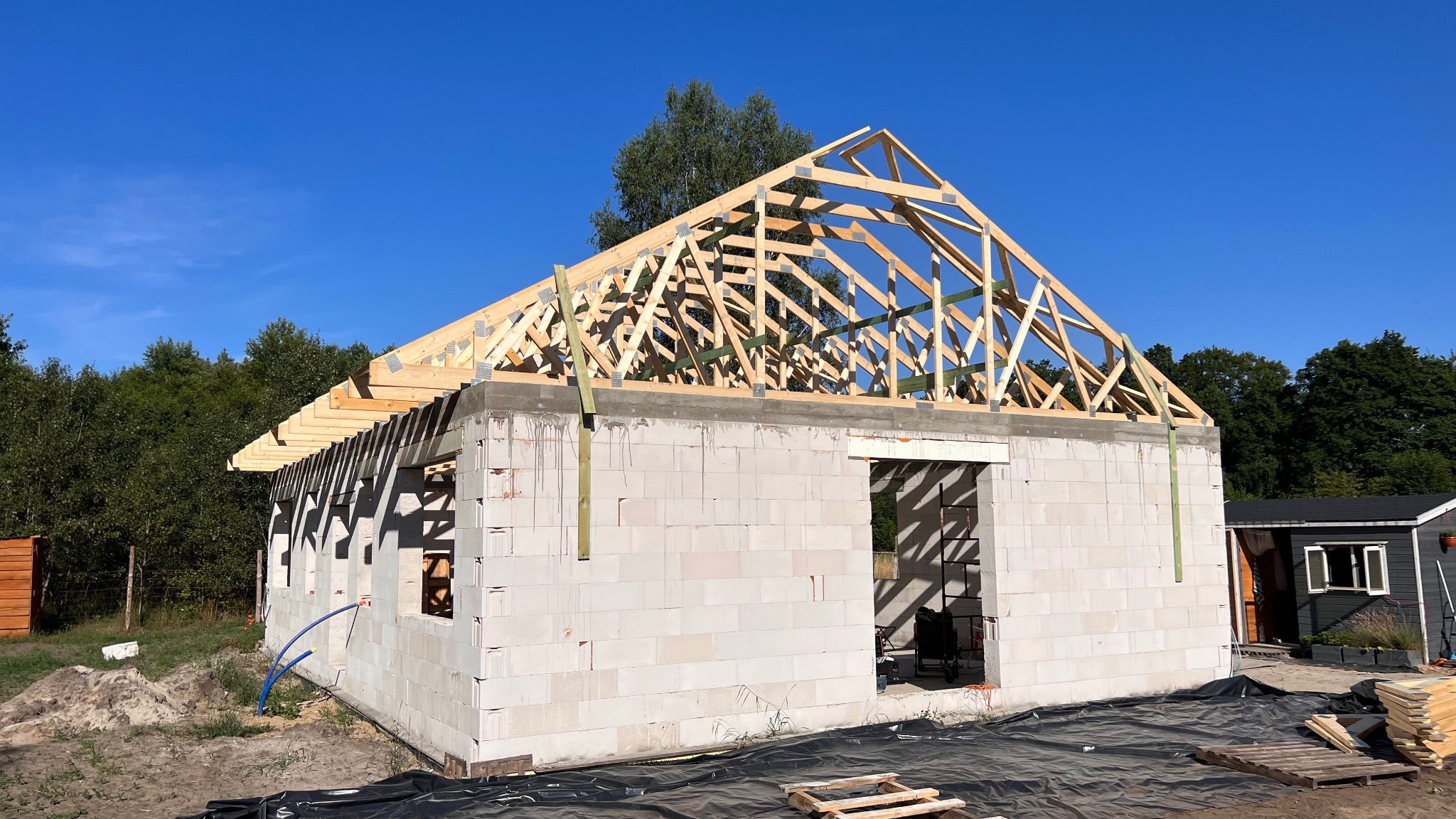 Budowa domu jednorodzinnego: ściany z betonu komórkowego i drewniana konstrukcja dachu w trakcie montażu, widok z zewnątrz w słoneczny dzień.