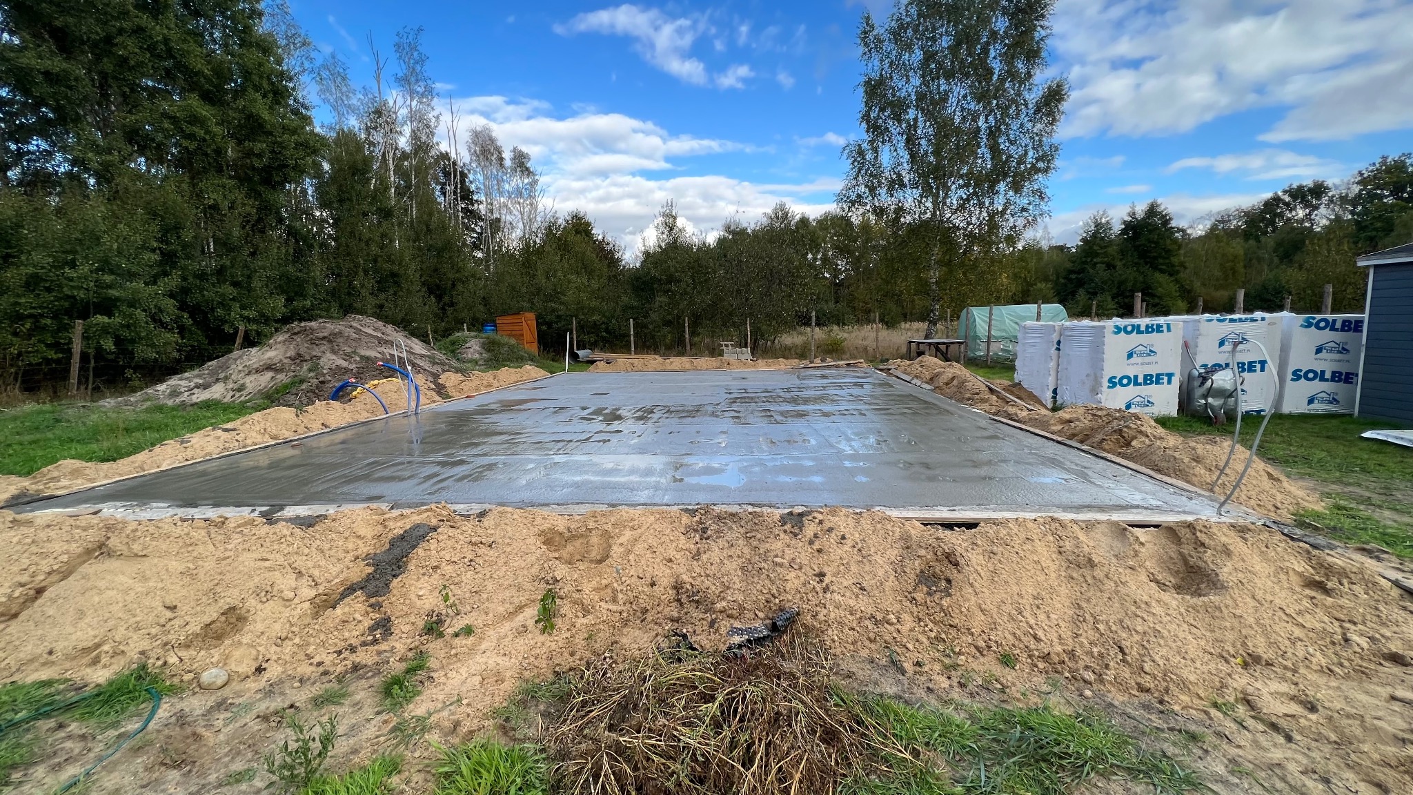 Świeżo wylana, mokra płyta fundamentowa na gruncie, otoczona piaskiem i deskowaniem. W tle składowane bloczki Solbet i zieleń drzew.