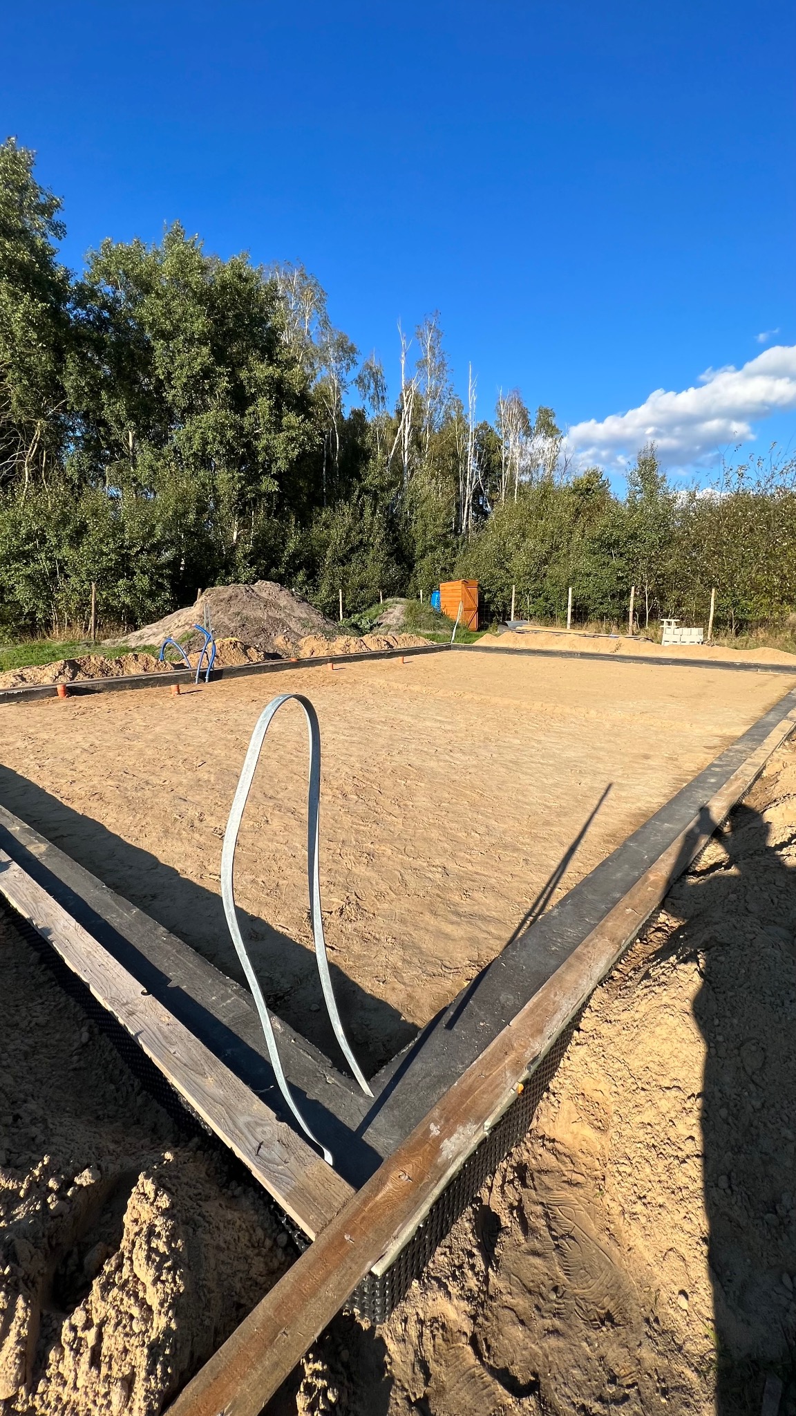 Przygotowany teren pod fundamenty domu z widocznym szalunkiem i izolacją, na tle zieleni i błękitnego nieba. Widoczny cień osoby.