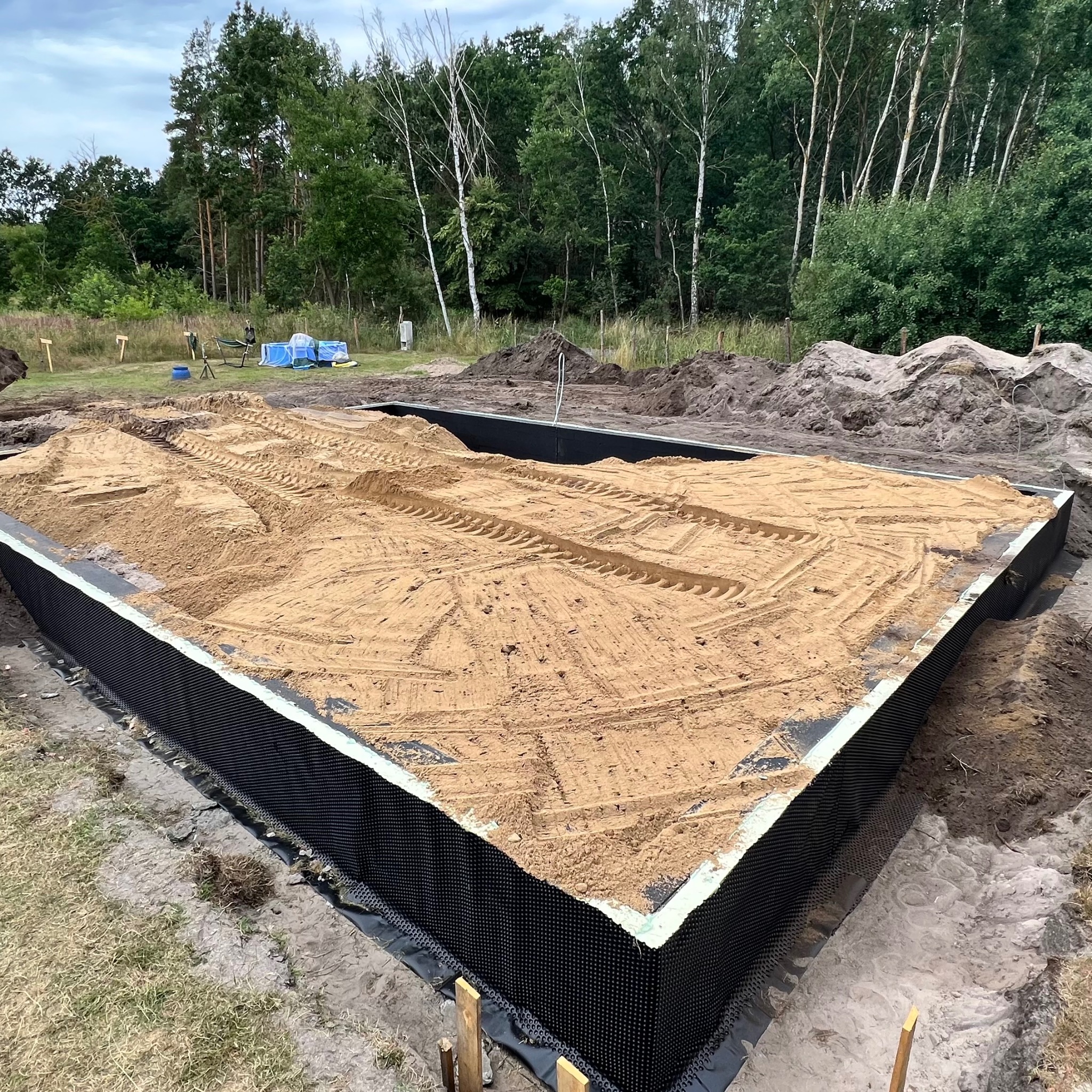 Przygotowany wykop pod fundament z izolacją pionową i zasypany piaskiem, widoczne ślady opon. W tle drzewa i sprzęt budowlany.