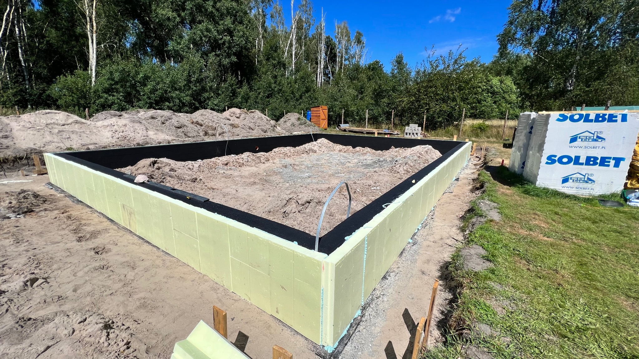Ocieplone fundamenty domu jednorodzinnego z widocznymi bloczkami Solbet w tle, na tle zieleni i błękitnego nieba. Etap budowy w słoneczny dzień.