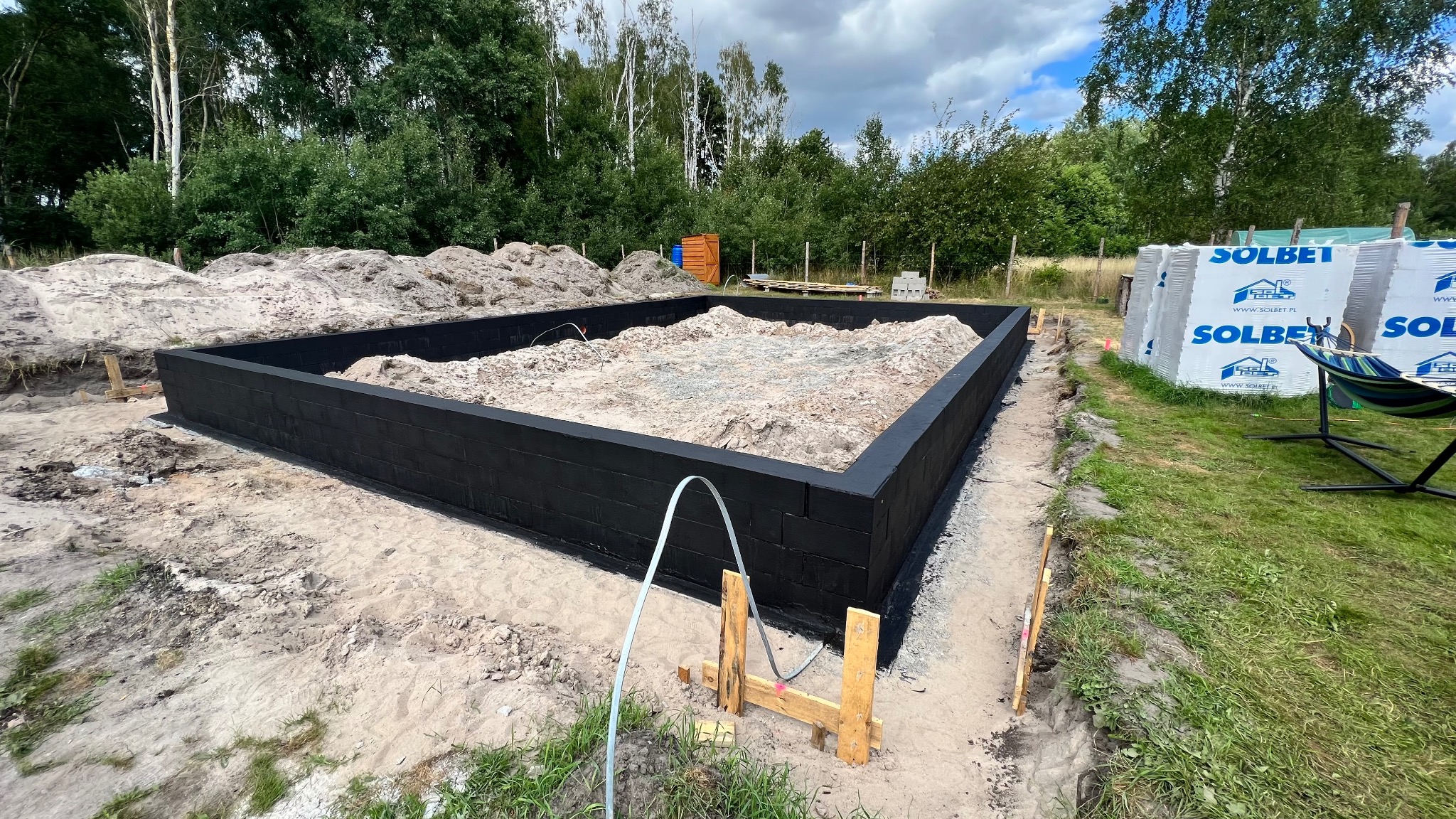 Zalane fundamenty z czarną izolacją, widok z góry na plac budowy. W tle drzewa i materiały budowlane Solbet. Etap budowy domu jednorodzinnego.