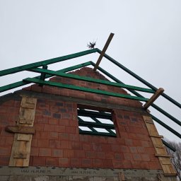 KMM BUDOWNICTWO SPÓŁKA Z OGRANICZONĄ ODPOWIEDZIALNOŚCIĄ - Wznoszenie ścian domu z czerwonej cegły, widoczne drewniane elementy konstrukcji dachu i szalunki okienne. Ujęcie z dołu, pochmurne niebo w tle.