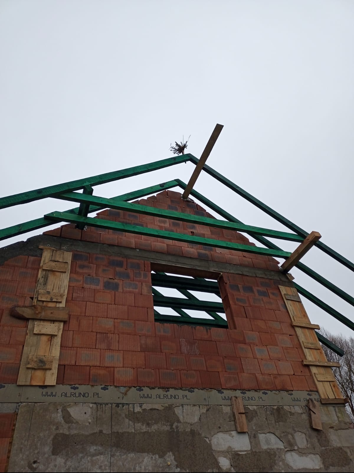 Wznoszenie ścian domu z czerwonej cegły, widoczne drewniane elementy konstrukcji dachu i szalunki okienne. Ujęcie z dołu, pochmurne niebo w tle.