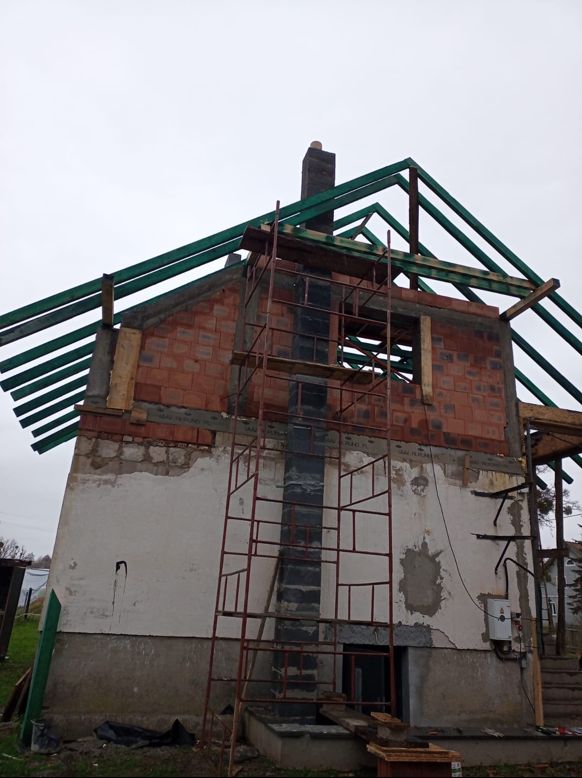 Budowa domu z czerwonej cegły, widoczne rusztowanie i elementy konstrukcji dachu. Częściowo otynkowana ściana z widocznymi uszkodzeniami. Ujęcie z dołu, pochmurne niebo.