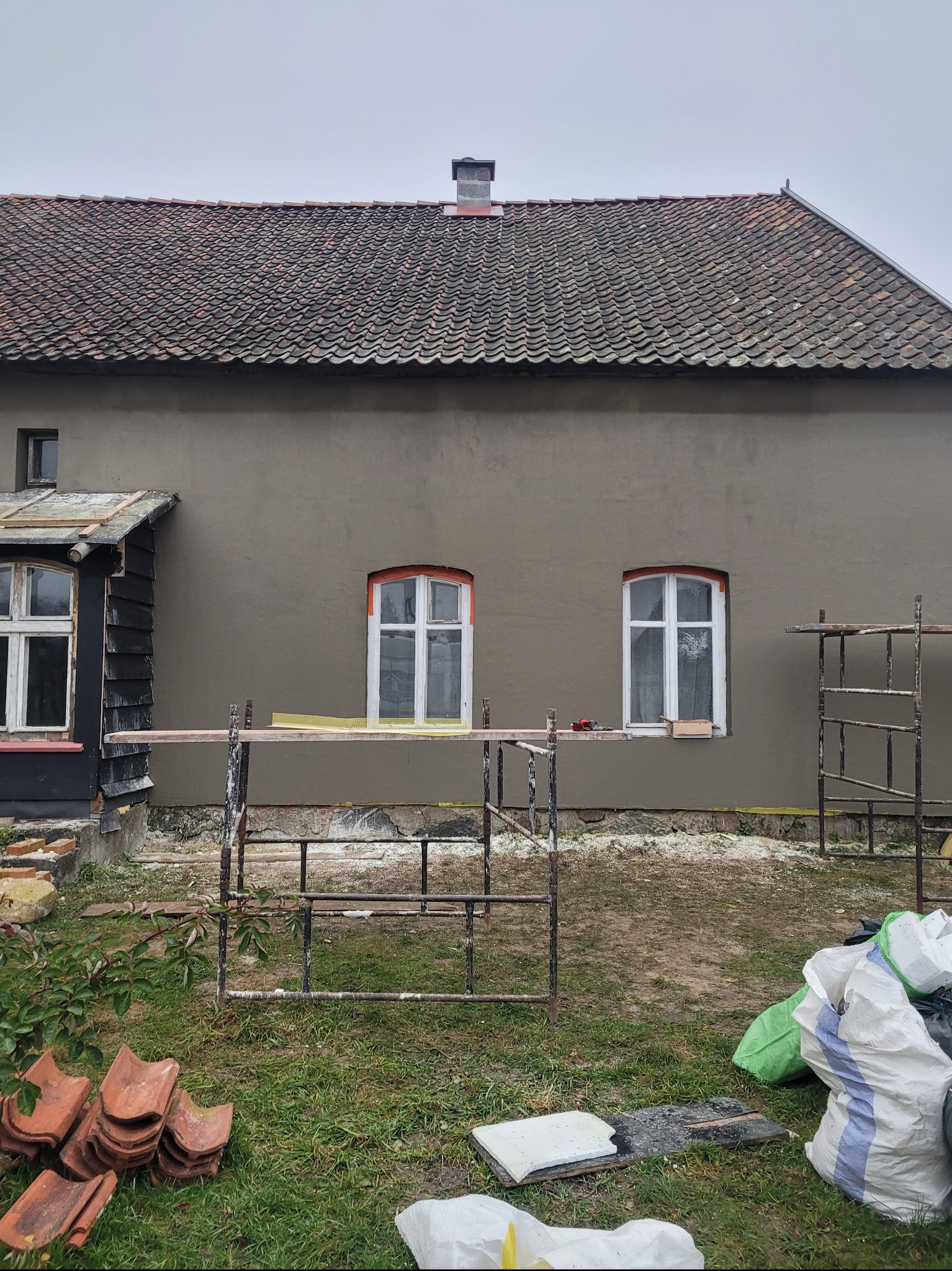 Elewacja domu w trakcie prac: świeżo nałożony tynk w szarym odcieniu, widoczne rusztowania, okna z białymi ramami i stary dach z dachówki ceramicznej.