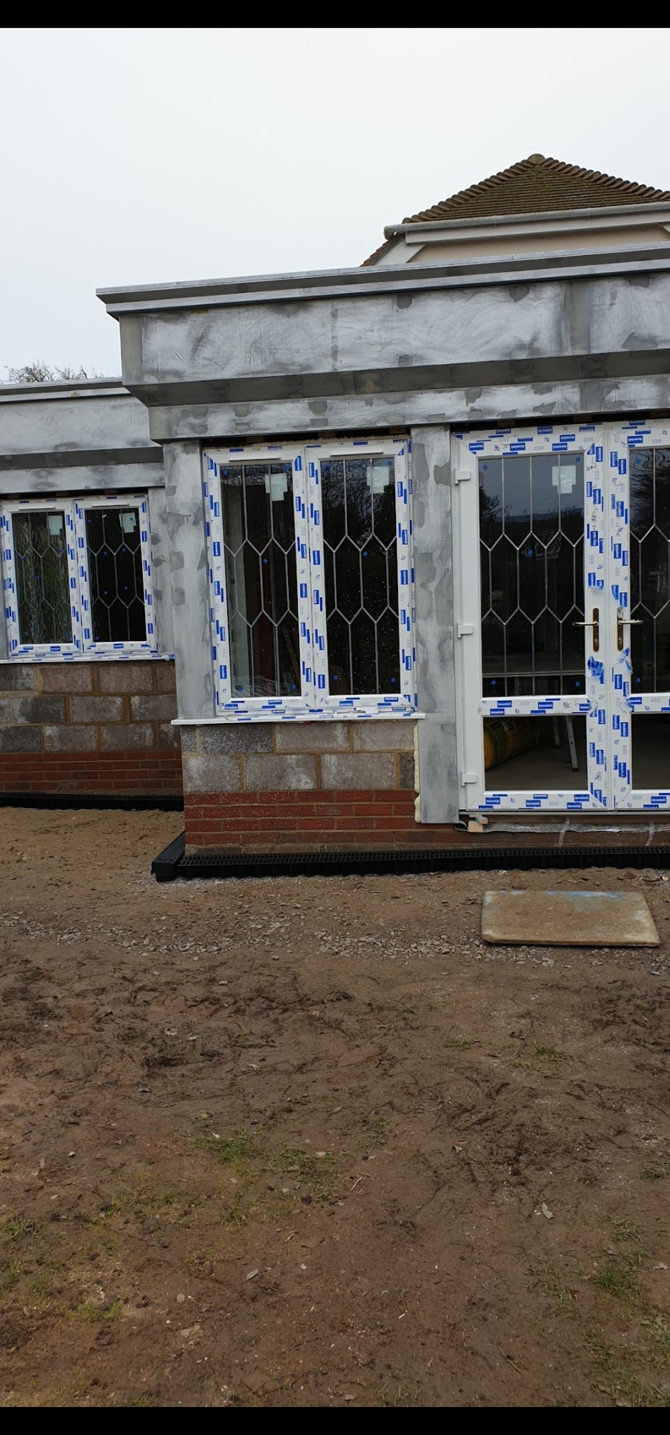 Budynek w trakcie wykończenia z oknami z białymi ramami i ceglaną podstawą. Ściany w stanie surowym, przygotowane do dalszych prac wykończeniowych. Widoczne elementy drenażu.