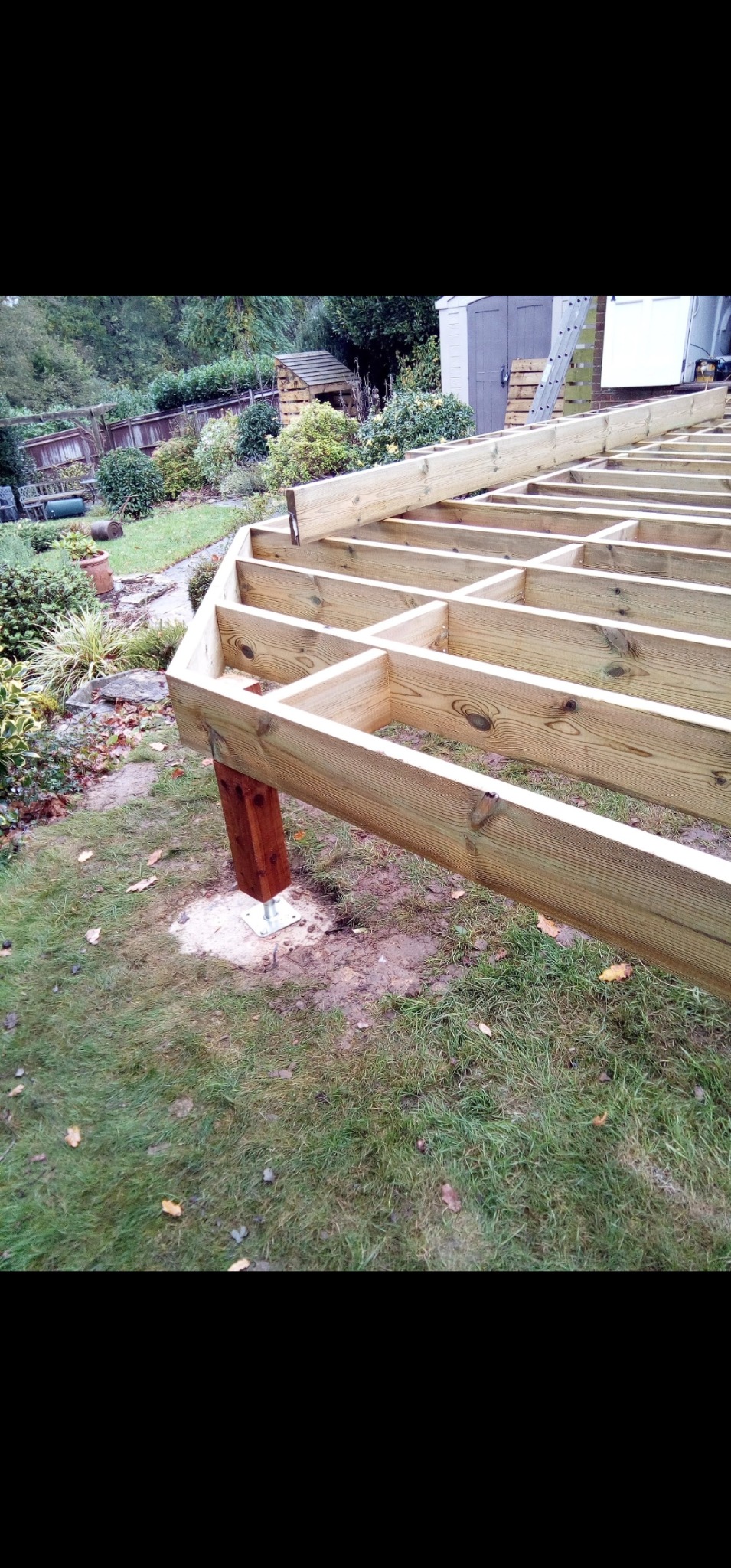 Drewniana konstrukcja tarasu w ogrodzie, widoczne legary i słupy na metalowych podstawach. W tle zieleń i budynki gospodarcze.