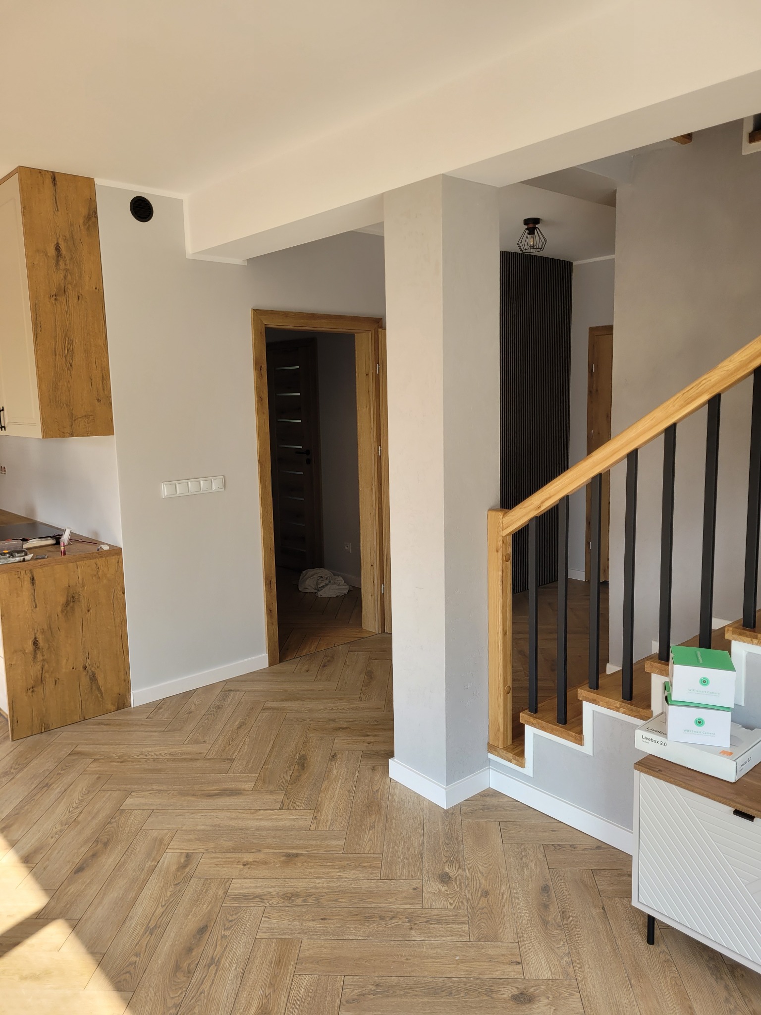 Jasne wnętrze domu po remoncie: parkiet w jodełkę, schody z drewnianą balustradą i szarymi ścianami. Widoczne elementy wykończenia kuchni i drzwi.