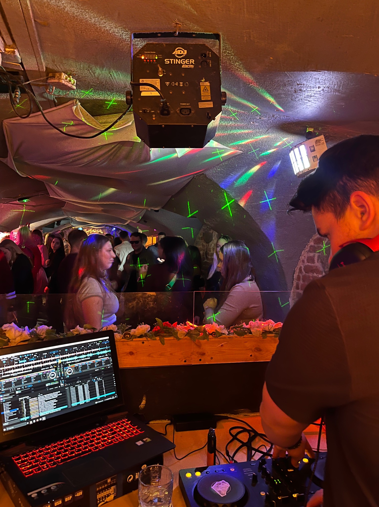 DJ z laptopem i konsoletą na tle bawiących się osób, oświetlonych kolorowymi światłami dyskotekowymi. Widoczny element dekoracji z kwiatów.