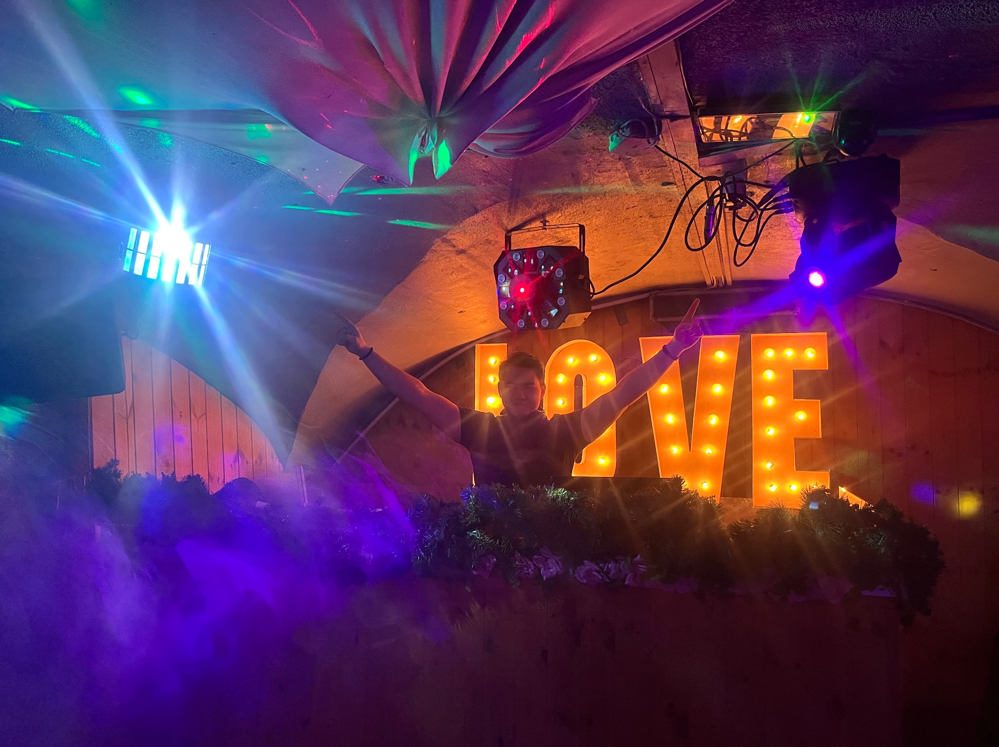 DJ z rozłożonymi rękami na tle podświetlonego napisu LOVE i dekoracji roślinnych, oświetlony kolorowymi światłami dyskotekowymi.
