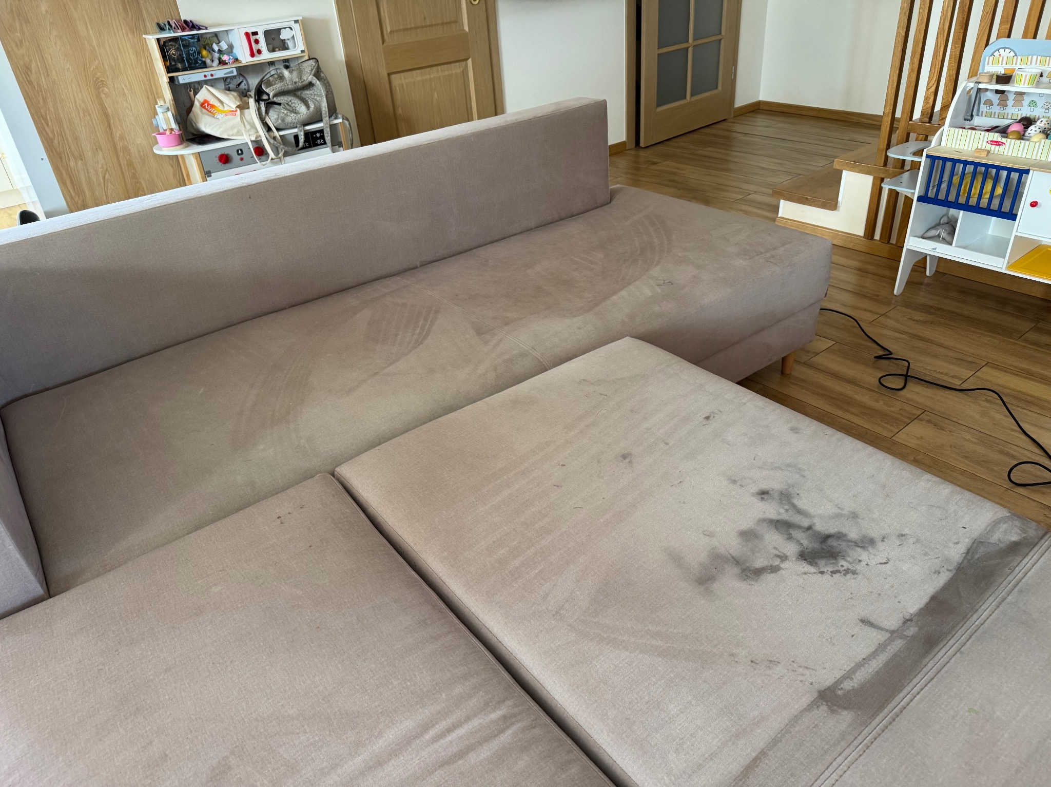 Brudna, beżowa kanapa modułowa z widocznymi plamami i śladami po czyszczeniu. W tle zabawki dziecięce i drewniana podłoga.