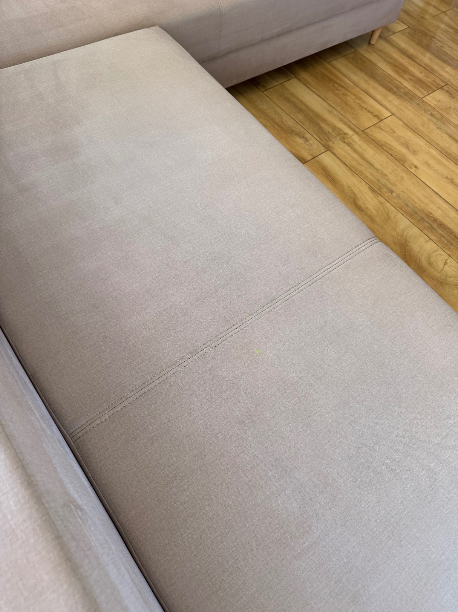 Jasna, beżowa kanapa z widocznym szwem i małą plamką na siedzisku, ustawiona na drewnianej podłodze. Ujęcie z góry, bliski kadr na fragment mebla.