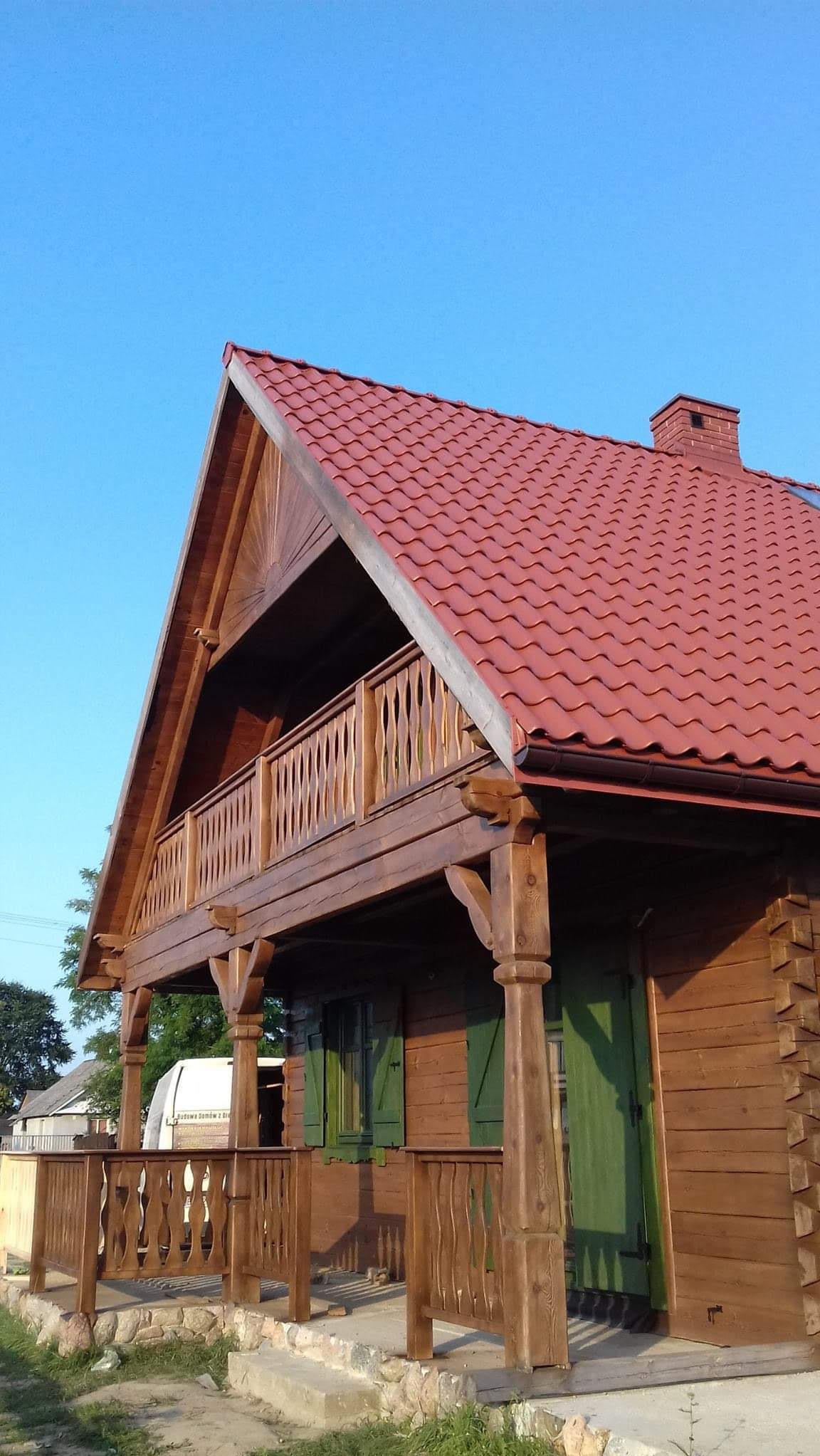 Drewniany dom z czerwonym dachem, balkonem i zielonymi okiennicami, widziany z dołu pod kątem. Charakterystyczne zdobienia balustrady i kolumn.