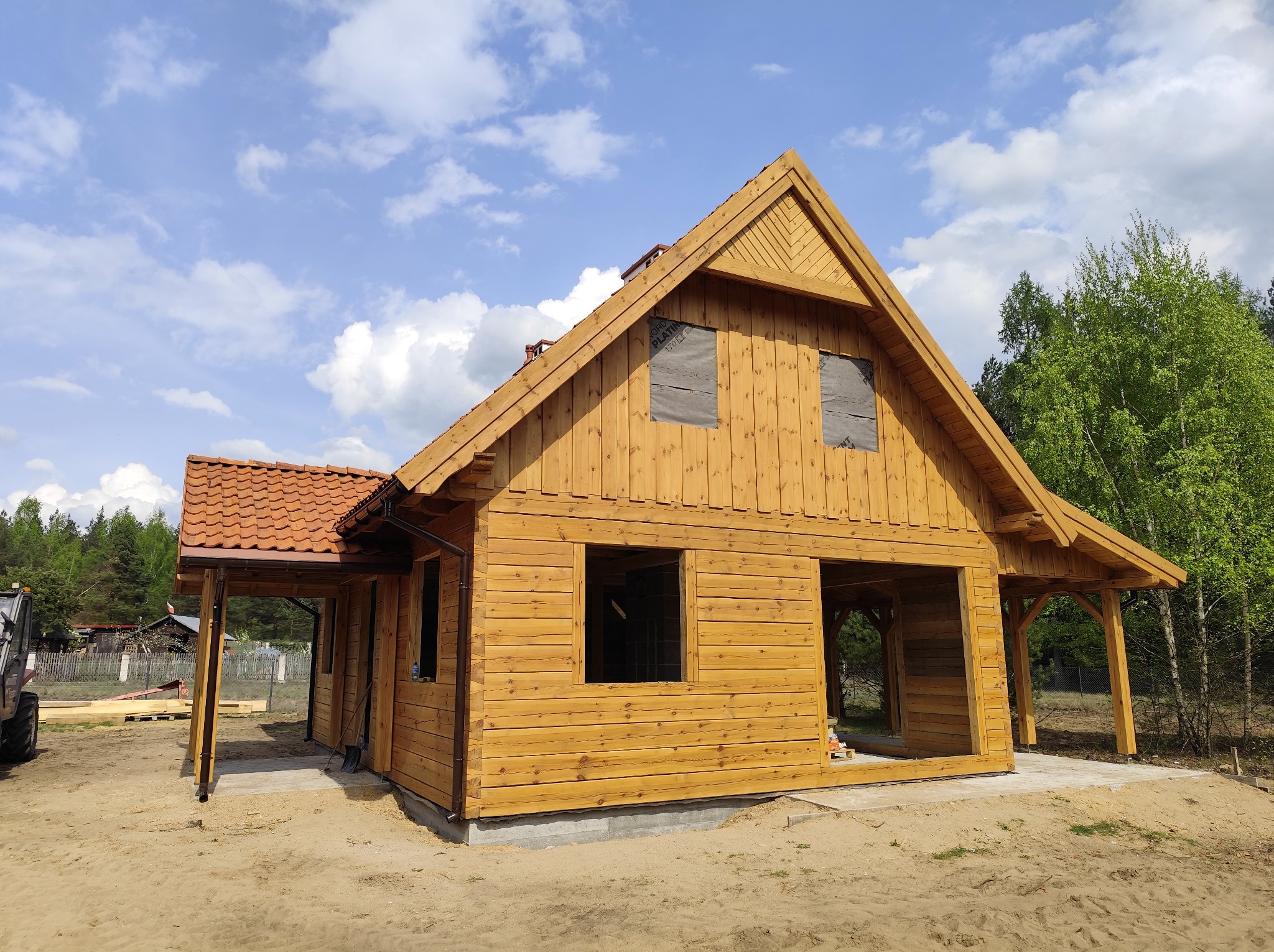 Surowy, drewniany dom z bali w trakcie budowy, z widoczną konstrukcją dachu i częściowo zamontowaną stolarką okienną. Czerwona dachówka, błękitne niebo w tle.