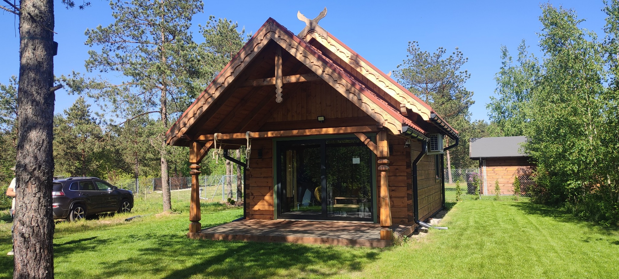 Drewniany domek letniskowy z przeszkloną ścianą frontową, zdobionym dachem i kolumnami, otoczony zielenią. W tle widoczny drugi budynek i samochód.
