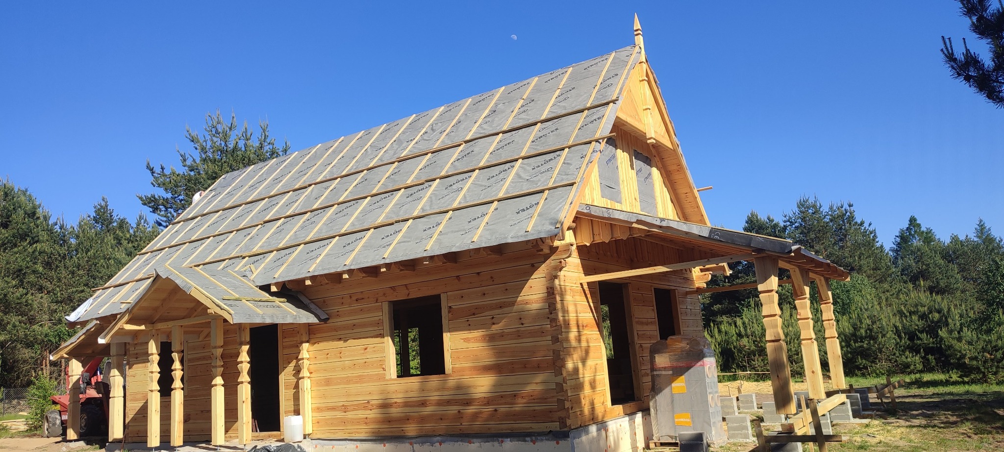 Surowy, drewniany dom z bali w trakcie budowy, z widoczną membraną dachową i elementami konstrukcyjnymi, na tle błękitnego nieba i zieleni drzew.