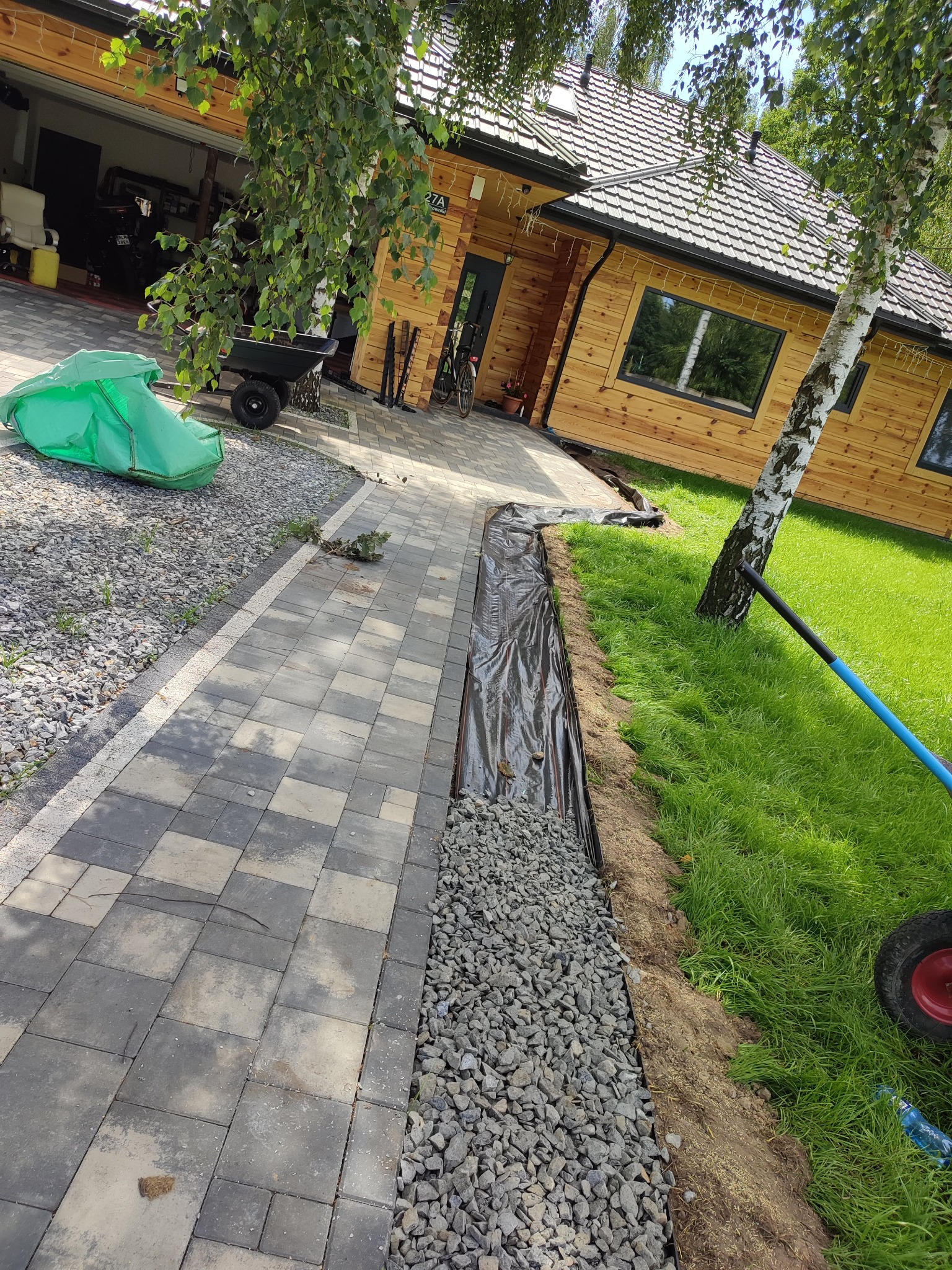 Układanie kostki brukowej i drenażu przy drewnianym domu z jasną elewacją. Widoczny fragment trawnika i drzewo. Prace w toku, z użyciem folii i kamieni.